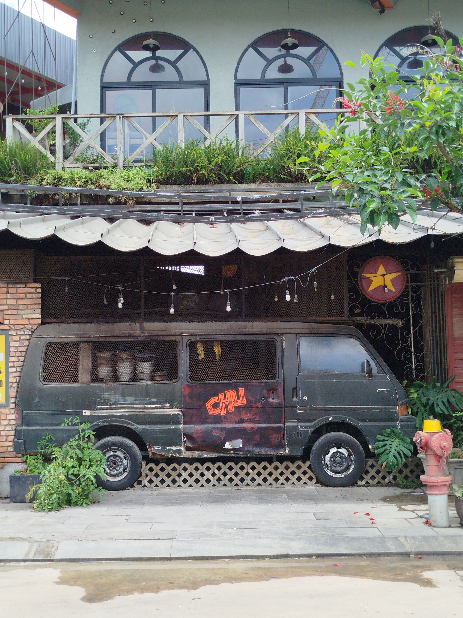 A dilapidated dark green van with the word "CHU" painted on the passenger door is parked outside a building in Da Lat, Vietnam. The van has rusted patches, a grill over the windows, and what appear to be bowls inside. The building has a canopy with scalloped edges and hanging light bulbs. Above the canopy is a balcony with a wooden railing and potted plants. The building’s second story has three arched windows. To the right of the van is a red and yellow fire hydrant and tropical plants. A sign with a yellow star is mounted on an ornate metal screen to the right of the van. The sidewalk in front of the van is wet.