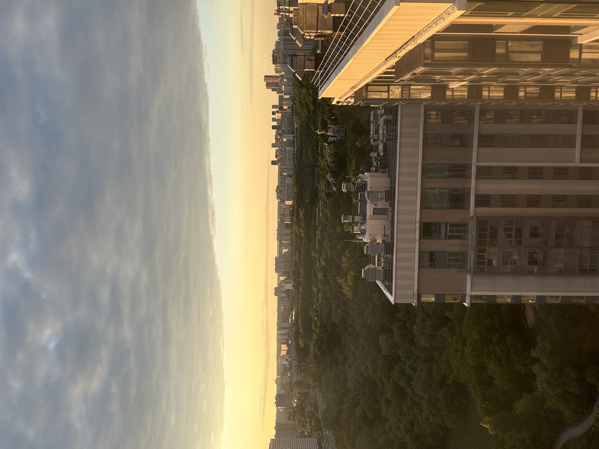 This media file is a vertical, slightly tilted photograph of a cityscape at dusk. The scene is dominated by a modern, multi-story building in the foreground, with its facade featuring numerous windows and external air conditioning units. To the left of the building, a vast expanse of clouds stretches across the sky, reflecting the warm hues of the setting sun.
Beyond the immediate building, a dense, dark green forest covers the landscape, interspersed with a variety of other buildings, some of which appear to be older high-rise apartment blocks. The setting sun casts a soft, golden glow over the horizon, creating a hazy atmosphere that blends the sky with the distant cityscape. A winding path is visible through the trees in the lower right portion of the image.
The time of day is clearly dusk or early evening, given the low angle of the sun and the warm, diffused light. The weather appears calm, with the clouds suggesting a peaceful transition from day to night. The presence of dense trees and the style of the buildings suggest an urban environment bordering a park or natural reserve. There are no people or discernible activities captured in this image, making it a landscape-focused shot. The overall mood is tranquil and somewhat serene, conveyed through the soft lighting and the quietude of the scene. No visible text is present in the image. The location context provided is Dolgoprudny, Russia.