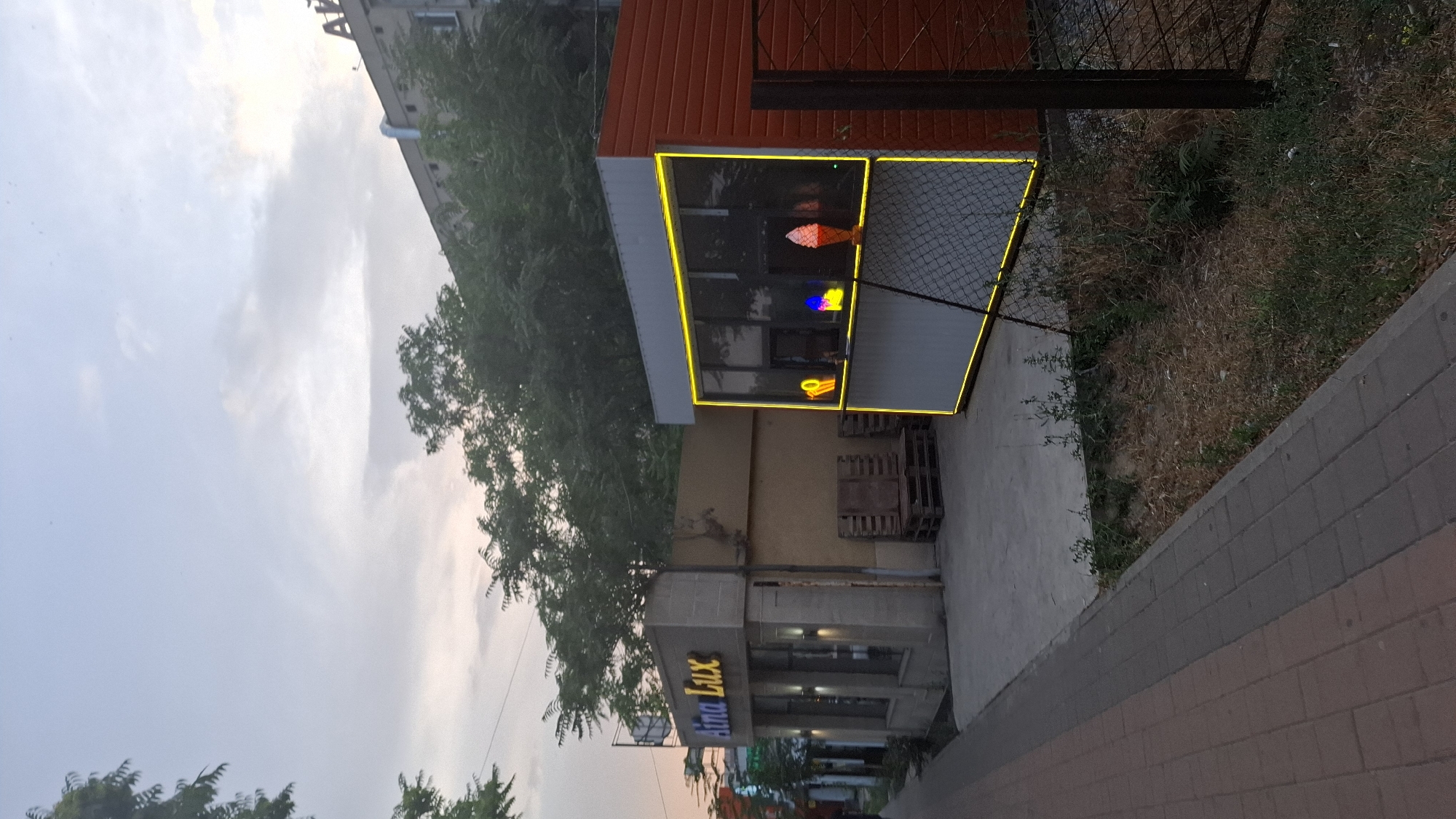 The image shows a street scene in Shymkent, Kazakhstan. The foreground features a paved walkway made of grey and reddish-brown bricks. To the right, a patch of dry grass and some greenery is visible, bordered by a chain-link fence. Beyond the fence, a small commercial building with a red-orange facade and white trim is illuminated by bright yellow neon lights outlining its windows. Inside the windows, colored lights are visible, resembling ice cream cones. Pallets are stacked near the building.
The middle ground shows another part of the commercial building with signage that reads "Extra Lux" in blue neon letters. This section also has illuminated windows. A large tree with dark green foliage partially obscures the view of the building. In the background, a multi-story building with a light-colored facade can be seen. The sky above is overcast with soft, diffused light, suggesting it is either early morning or late evening. The overall impression is a slightly rundown urban environment with a small business operating. No people are visible in the frame.