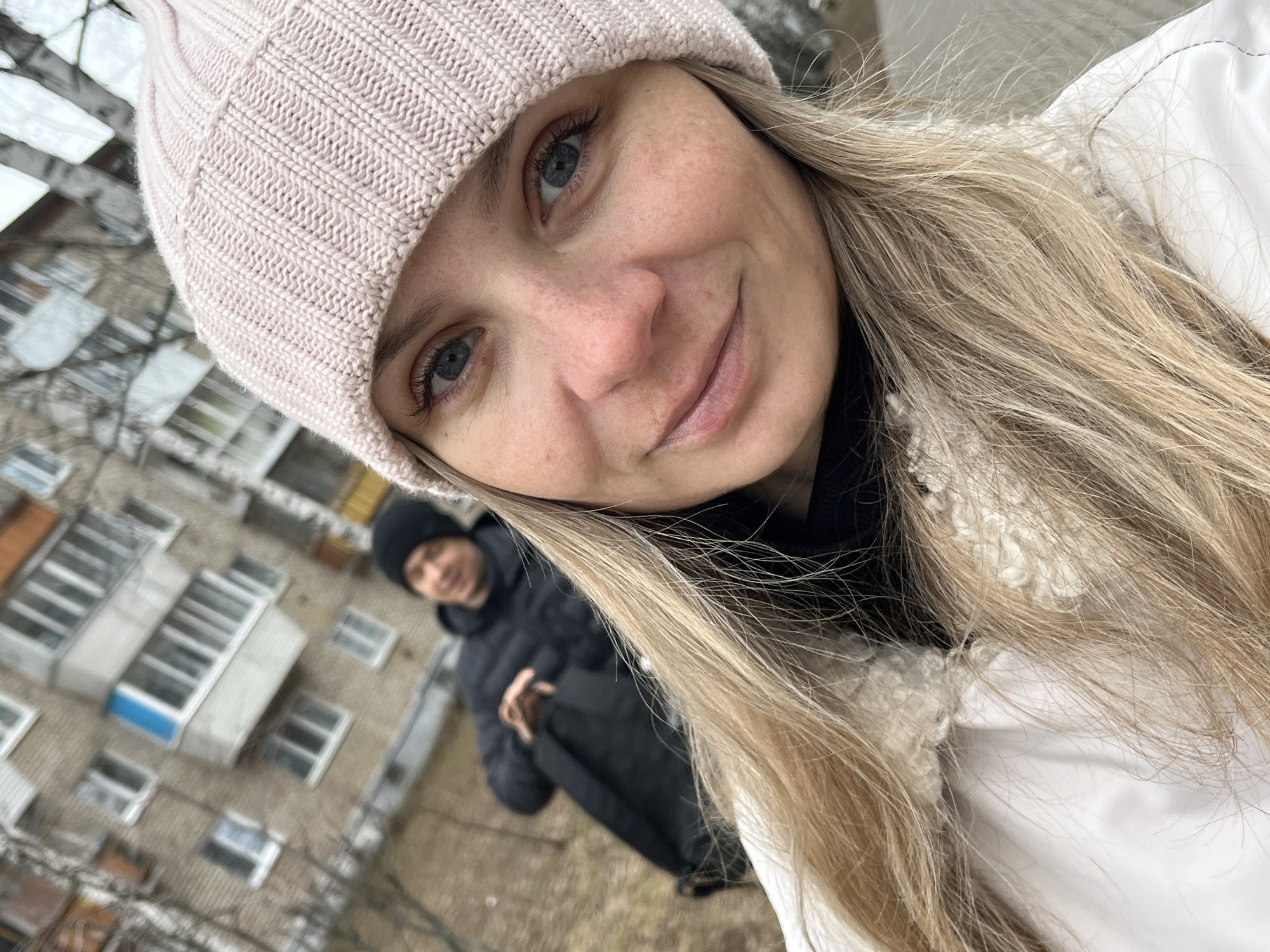 A woman with blonde hair and blue eyes wears a pink beanie and a white jacket with a furry collar. She is looking directly at the camera with a slight smile. In the background, out of focus, a man in a black jacket and beanie stands on an overcast day in front of a gray apartment building. The building appears to be in Chaikovsky, Russia, based on the architectural style typical of that region.