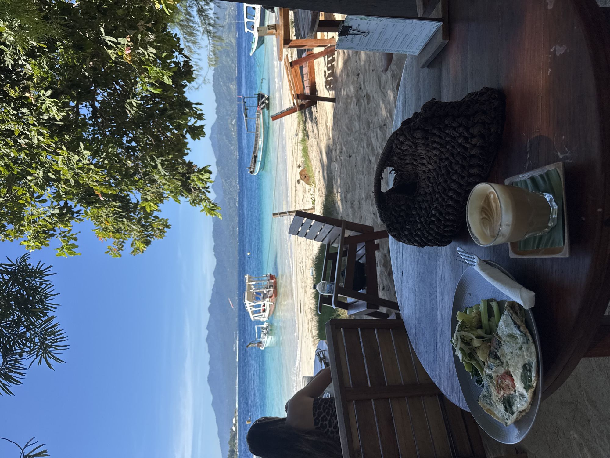 A sunlit scene depicts a partially obscured outdoor dining area overlooking a beach in an unknown Indonesian location. A round, dark wooden table dominates the foreground, adorned with a plate containing what appears to be an omelet and a side of vegetables, a fork wrapped in a white napkin, a glass of coffee sitting on a small woven coaster, and a dark woven bag.
Beyond the table, the beach stretches into the distance, displaying several wooden boats anchored in the turquoise water. Light wooden benches and tables are scattered on the sand, suggesting a relaxed atmosphere. A woman with dark hair and wearing a patterned top is partially visible in the lower right corner of the image.
In the background, a mountain range fades into the clear blue sky, adding depth to the scene. Green foliage from a tree hangs over the left side of the image, providing a natural frame.
