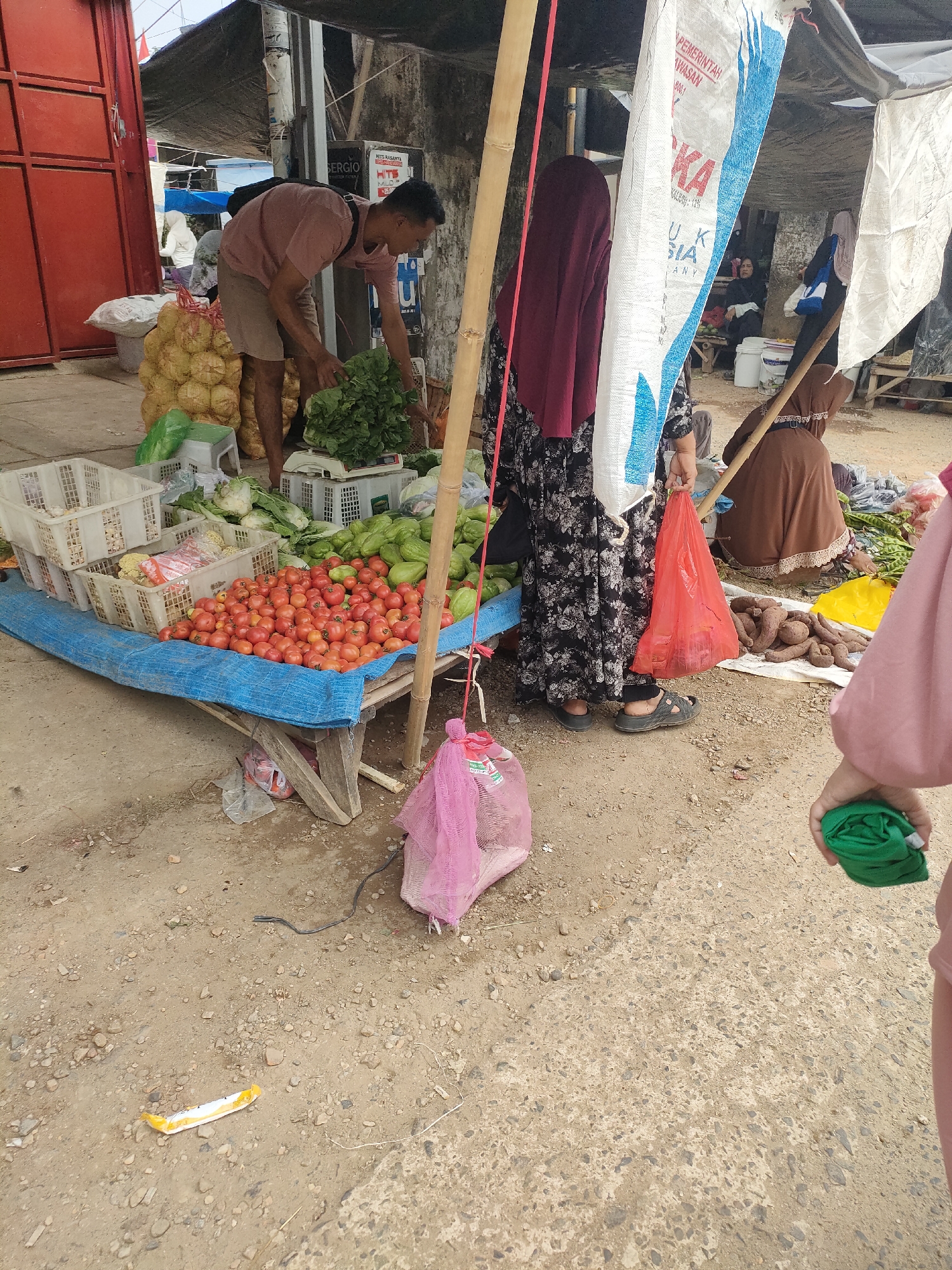 The image captures a vibrant scene at an outdoor market in Sukoyoso, Indonesia, likely on a warm, cloudy day. In the foreground, a variety of fresh produce is displayed on a wooden stall covered with a blue tarp. Piles of red tomatoes are prominent, alongside baskets of green chayote squash and other leafy vegetables. Two large sacks of yellow-skinned coconuts are stacked near the stall.
Several individuals are present, engaged in market activities. A man in a pink t-shirt and shorts is seen bending over a scale, seemingly weighing vegetables. A woman wearing a dark floral dress and a burgundy hijab stands near the stall, holding a red plastic bag. Another woman, in a brown hijab and dress, is seated behind a pile of sweet potatoes, some of which are visible on a white sheet. A person in a light pink garment is partially visible on the far right, holding a rolled-up green fabric.
The market appears to be set up under a makeshift shelter, with tarpaulins and bamboo poles providing shade. Bags and miscellaneous items are scattered on the ground, suggesting a bustling and active marketplace. The ground is a mix of dirt and gravel. In the background, more stalls and structures are visible, hinting at the larger scale of the market. A large red metal door is present to the left of the scene. A white plastic bag with blue and red lettering, possibly advertising, hangs prominently from a bamboo pole in the midground.