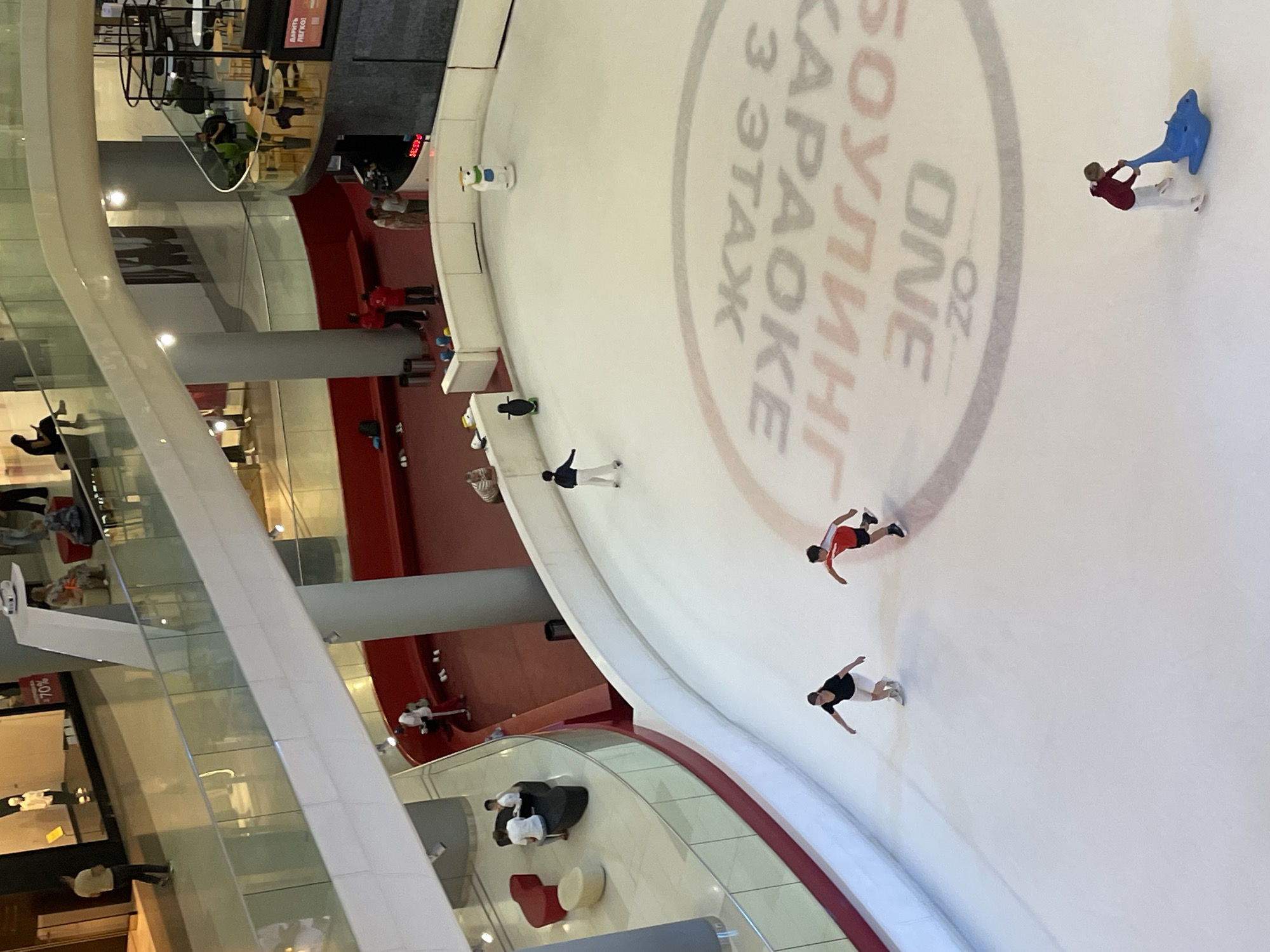 The image shows an indoor ice skating rink located in the OZ Mall in Krasnodar, Russia. Several people are visible on the ice, including a woman in a black shirt and white shorts, a boy in a red and white shirt, and a person in white pants and a dark jacket. There are also several decorative figures on the ice, including a penguin and a polar bear, as well as a blue dolphin-shaped sled. The rink has a circular design with Cyrillic text printed on the ice surface. In the background, the mall's architecture is visible, including glass railings, seating areas, and storefronts. The overall scene depicts a recreational activity taking place within a commercial setting.