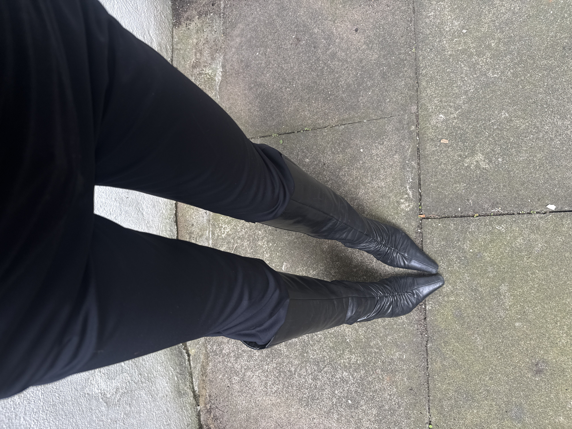 A low-angle shot depicts a person wearing black clothing and black pointed knee-high boots standing on a concrete surface. The person is angled towards the left, with their legs slightly apart. The concrete surface appears to be part of a sidewalk or paved area, showing signs of weathering and discoloration. The absence of other subjects and environmental cues makes it impossible to determine the exact location in London.