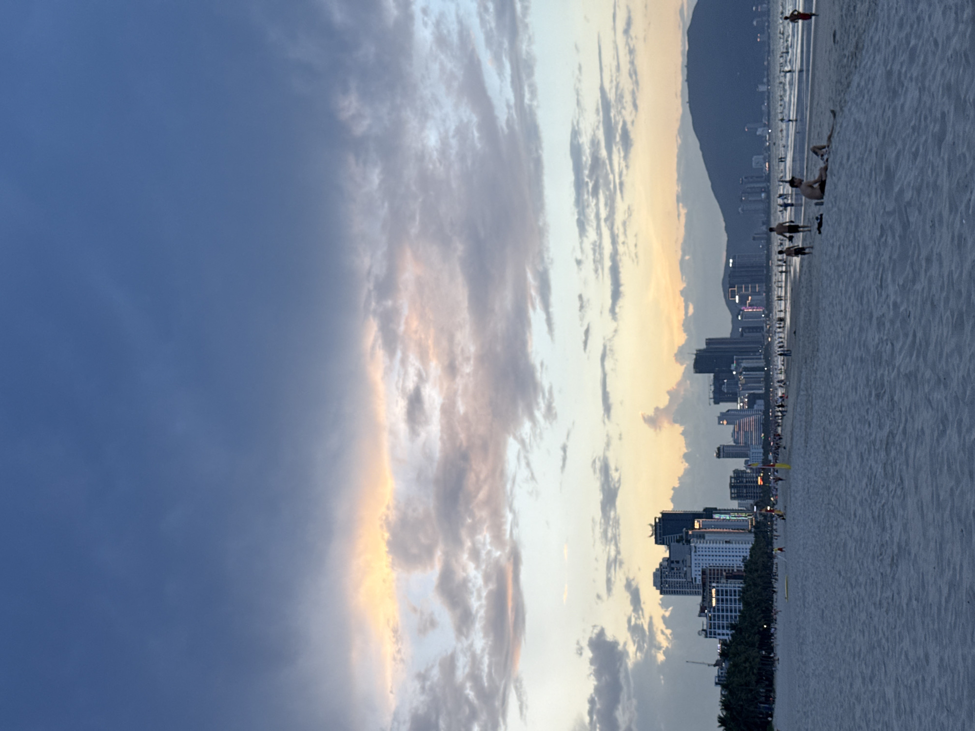 The image depicts a panoramic coastal scene at what appears to be sunset or dusk. The sky dominates the upper portion, showcasing a dramatic blend of colors and cloud formations. On the left, the sky is a deep, muted blue, gradually transitioning to warmer golden and soft orange hues towards the right, where the setting sun's light catches the edges of streaky, elongated clouds. Some clouds are dark and voluminous, while others are lighter and more ethereal, creating a dynamic atmospheric display.
In the foreground, a wide, sandy beach stretches horizontally. Scattered along the beach are several individuals, mostly appearing as dark silhouettes or dimly lit figures. Some people are standing, possibly strolling near the water's edge, while others are seen sitting or reclining on the sand, enjoying the evening. The texture of the sand is visible, showing subtle ripples and footprints.
Behind the beach lies a modern city skyline. Numerous high-rise buildings, some illuminated with early evening lights, line the coastline. These structures vary in height and design, indicating a developed urban area. Further back, a large, dark mountain range forms the backdrop, its peaks silhouetted against the softer sky, completing the scenic view.
The overall activity is leisurely, with people relaxing on the beach during the transitional period of daylight. No specific event is apparent beyond general beach-going. The notable details include the striking sky, the blend of natural and urban landscapes, and the tranquil mood conveyed by the soft, fading light. No visible text is present in the image. The location context indicates this is likely Đà Nẵng, Vietnam, a city known for its scenic beaches and mountainous surroundings.