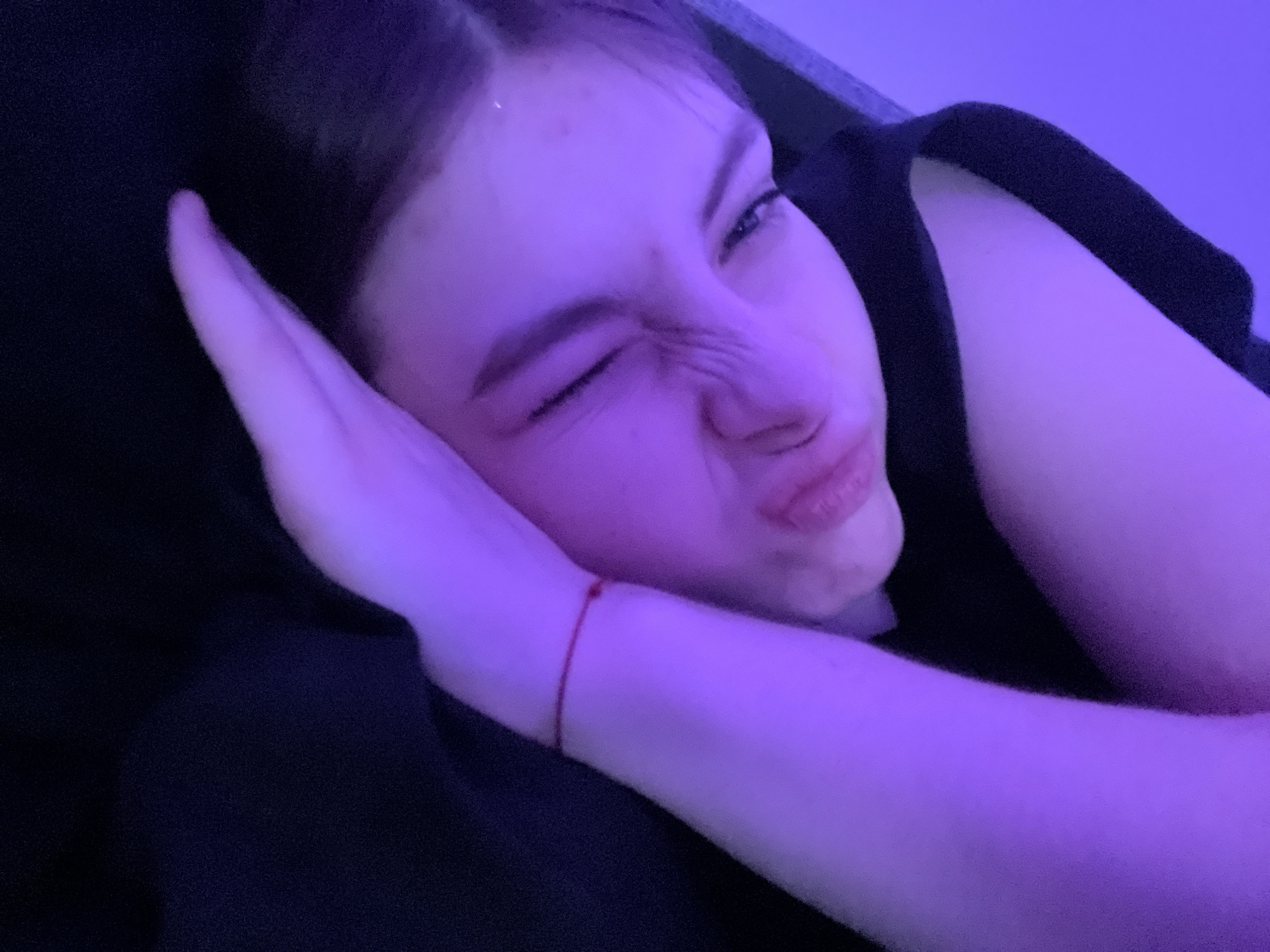 A young, fair-skinned woman is lying down, propped up on her arm and hand. Her face is contorted into a grimace, one eye is closed, and her lips are pursed. A red string is visible around her wrist. She is wearing a black top. The lighting is predominantly purple. There is no evidence in the image to indicate this image was taken in Saint Petersburg, Russia.