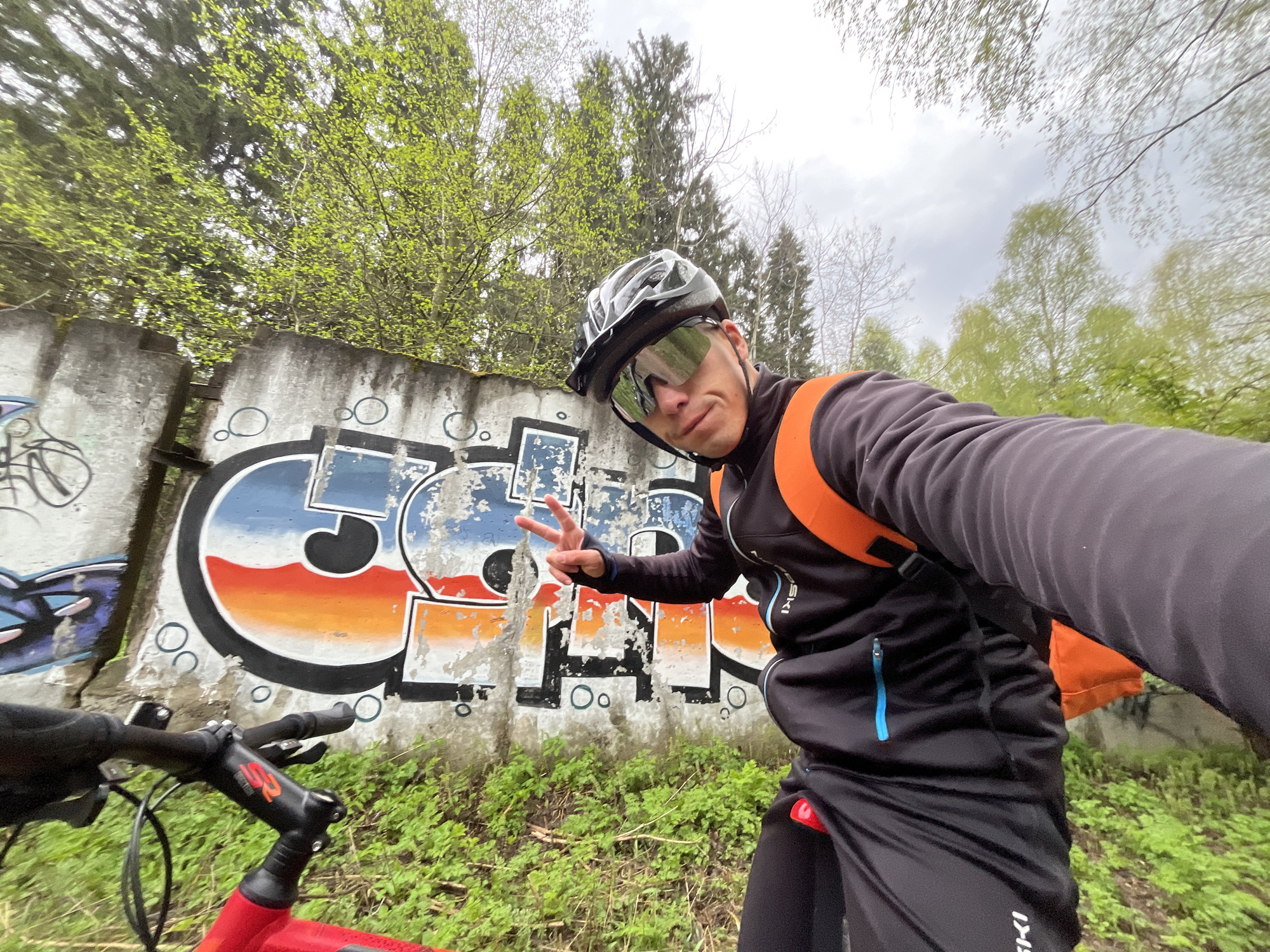 A man wearing a black cycling helmet and sunglasses is taking a selfie in front of a graffiti-covered concrete wall. He is wearing a black jacket with orange straps over his shoulders and making a peace sign with his left hand. The graffiti on the wall is colorful with a mix of blue, orange, and black. The handlebars of a red bicycle are visible in the bottom left corner. The area behind the wall has green grass and some trees with green leaves, and the sky is cloudy. The location is in Chaikovsky, Russia.