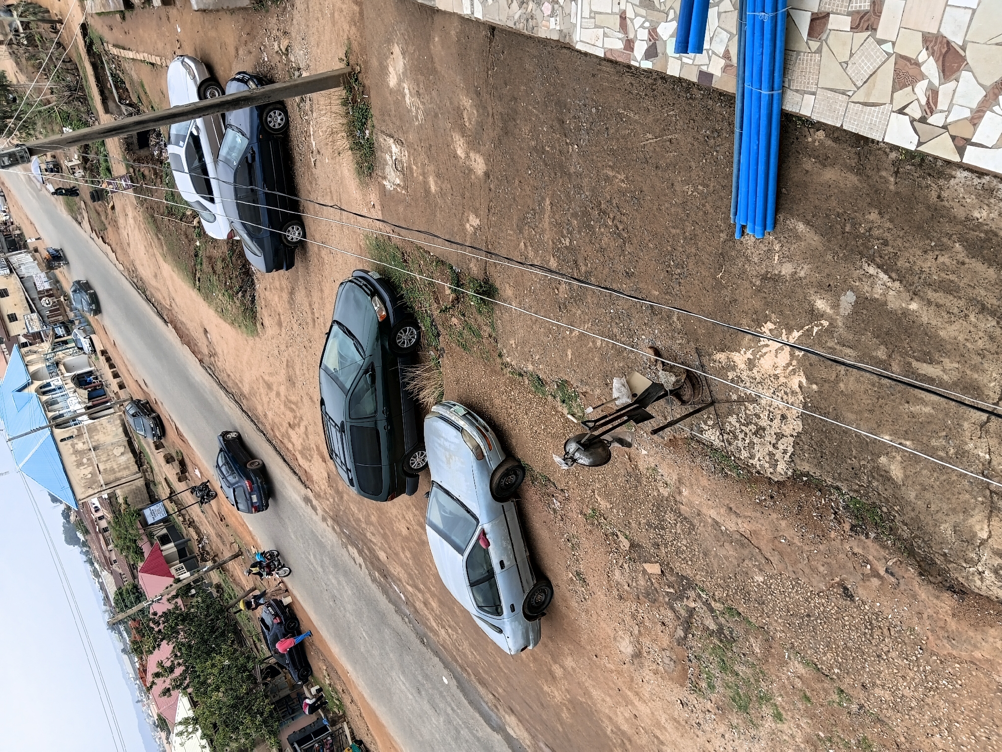 The image depicts a street scene in Jos, Nigeria, viewed from an elevated perspective. Several cars are parked along the street, with a mix of sedans and SUVs visible. The road appears paved, though with signs of wear. Adjacent to the road is an unpaved, dirt area. Buildings line the street, a mix of residential and commercial structures. Overhead power lines traverse the scene. A few pedestrians and motorcycles are present on the street. A bundle of blue pipes lies near a building with a mosaic-style facade.