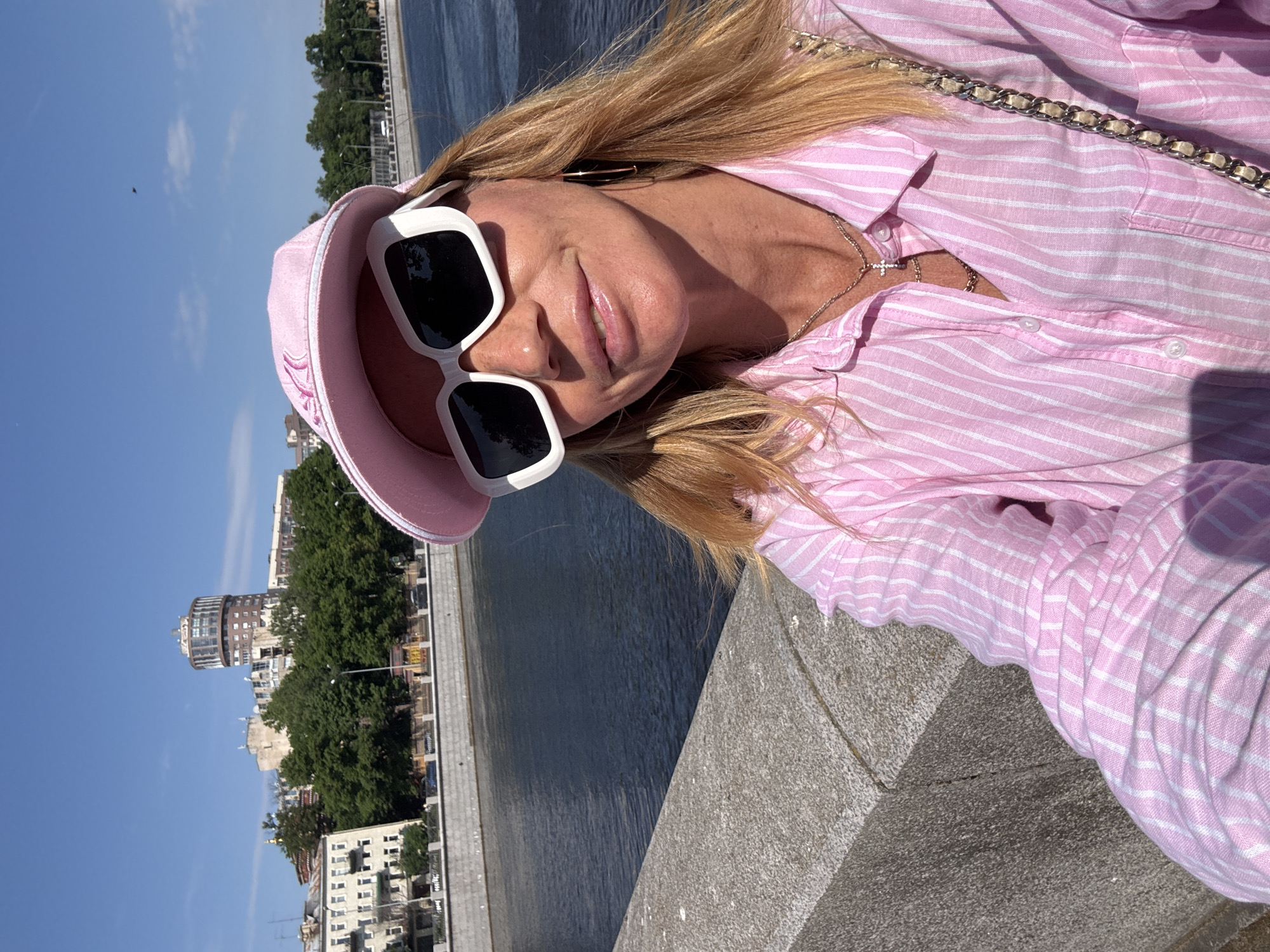 A blonde woman wearing a pink striped shirt, a pink baseball cap, and white rectangular sunglasses is taking a selfie on a sunny day. She is standing on a stone embankment overlooking a wide river. In the background, buildings and trees are visible. The architecture of the buildings suggests the location is likely Saint Petersburg, Russia, possibly near the Moskva River due to the style of the buildings and the presence of a tall, cylindrical building with a rounded top.