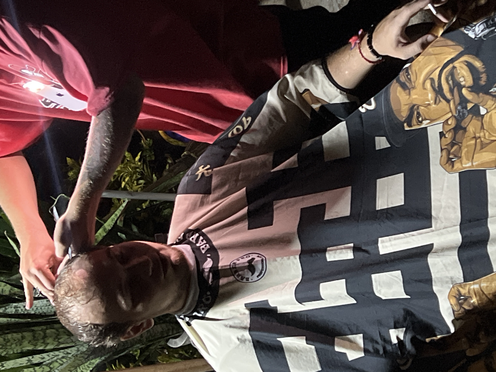 A person is receiving a haircut outdoors in an unknown location within the city of Unknown. The individual is seated with a patterned barber cape draped around their neck. A person with tattoos on their arm is standing and giving them a haircut. A hand holds a lit cigarette. Nearby are plants.