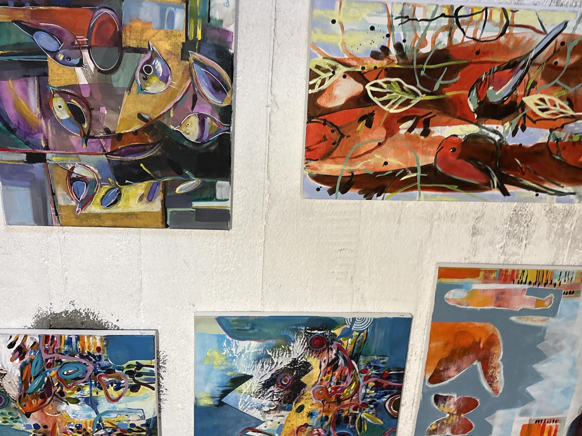 The image displays a collection of abstract paintings with a focus on birds and organic shapes, mounted on a textured white wall. The paintings are varied in size and orientation, creating a dynamic arrangement.
One prominent painting in the top right features a group of stylized birds against a backdrop of geometric shapes and a golden, textured area. The birds are rendered in vibrant colors with bold outlines.
To the left, a larger, vertical painting depicts an abstract scene with a central bird-like figure and various colorful elements, including swirling shapes and sharp, angular forms.
Below this, a horizontal painting shows two birds in a reddish-orange abstract environment with stylized leaves and branches.
Further down and to the left, another horizontal painting features a butterfly and a winged shape against a light blue background, with abstract elements of red, orange, and yellow.
The wall itself appears to be made of concrete blocks or a similar textured material, with visible lines and imperfections. The lighting suggests an indoor setting, possibly an art gallery or studio, with the natural light contributing to the scene. The overall impression is one of a curated display of modern, expressive artwork. The specific location within Gryllefjorden, Norway, is not discernible from the visual information provided in the image.