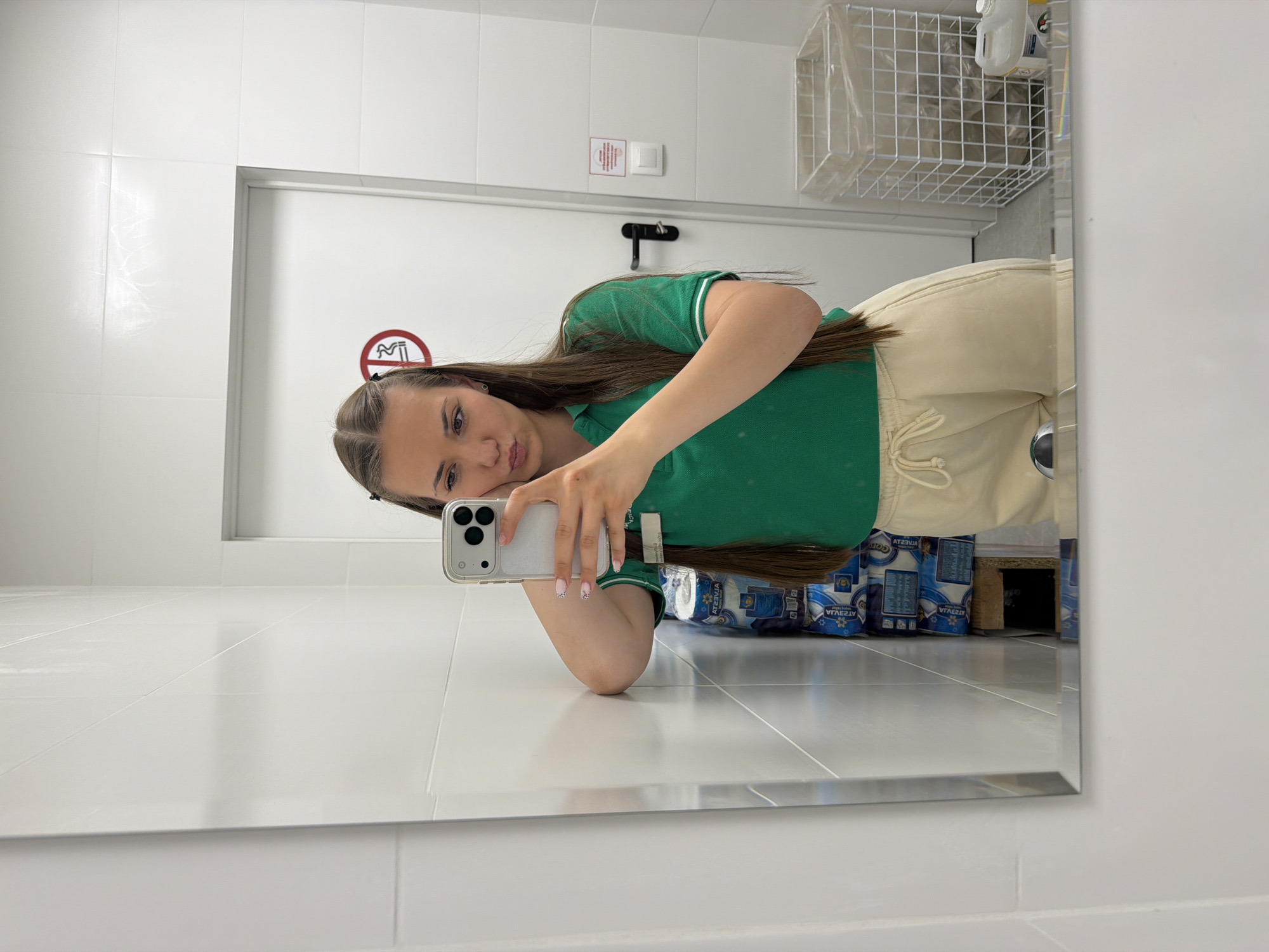 A young woman is taking a selfie in a bathroom mirror. She is wearing a green polo shirt and light-colored pants. Behind her, visible in the mirror, are stacks of toilet paper and a wire basket containing cleaning supplies. A "no smoking" sign is on the wall near the door. The bathroom appears to be in a public space, potentially a business or institutional building. There is no definitive information in the image to confirm the exact location in Sofia, Bulgaria.