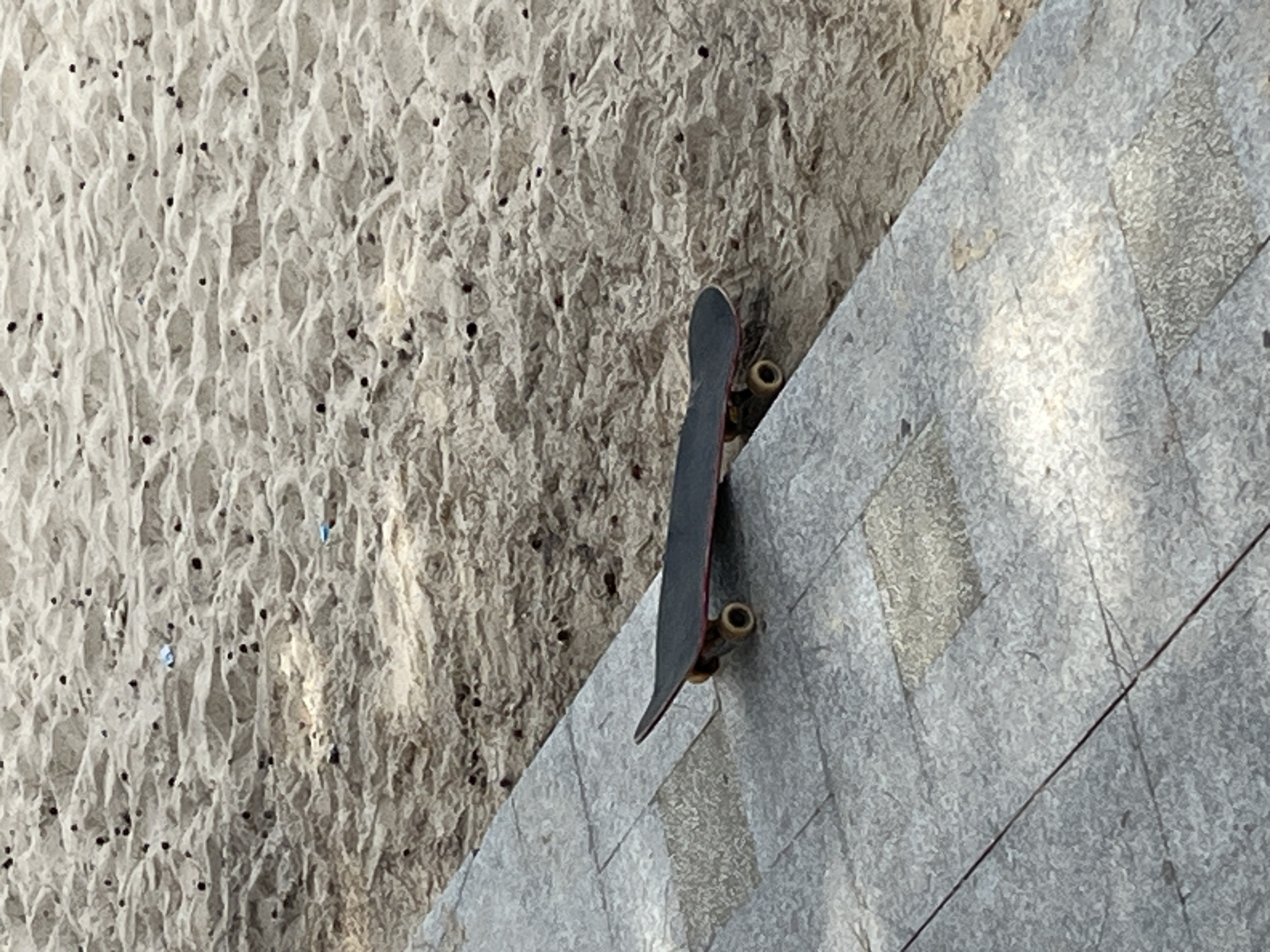 A black skateboard with red trim rests on a paved walkway next to a sandy embankment. The sand appears to be from a beach, with small dark objects scattered across its surface. The walkway is made of gray stone tiles. The scene is likely in Mũi Né, Vietnam, a popular tourist destination known for its beaches and sand dunes.