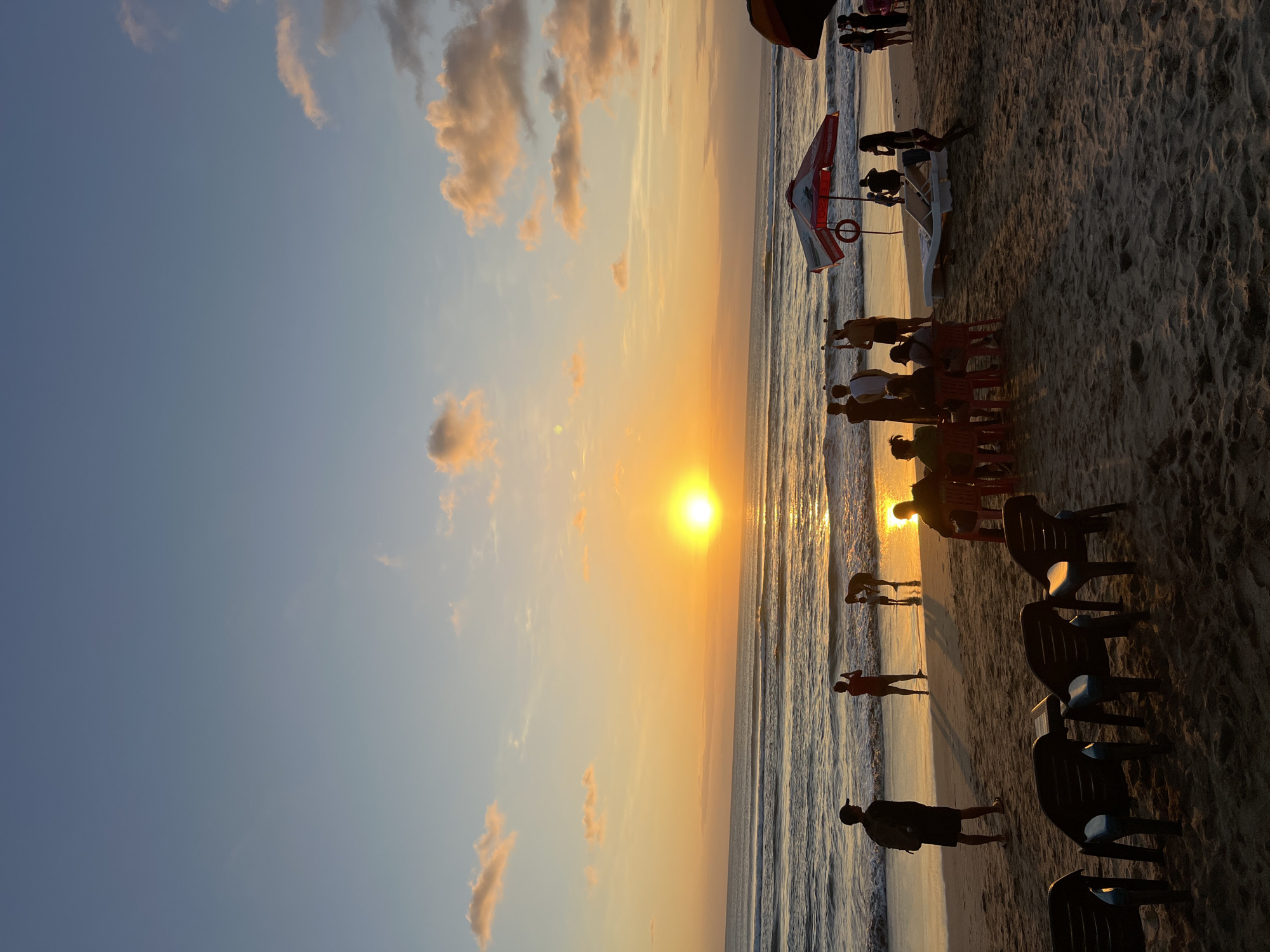 A sunset casts a warm orange glow over Kuta Beach in Indonesia. The sun is a bright orb on the horizon, reflecting off the gentle waves of the ocean. Silhouettes of people dot the sandy shore. Some are standing near the water's edge, perhaps enjoying the last rays of light or wading in the shallows. Others are gathered on the beach, some seated at tables with chairs, possibly dining or socializing. A red and white umbrella stands open, providing shade, with a life preserver hanging from it. Lounge chairs are also visible on the sand. The sky above transitions from a pale blue near the top to shades of orange and pink as it meets the horizon, with a few wispy clouds catching the fading light. The overall scene depicts a tranquil evening at a popular tourist destination.