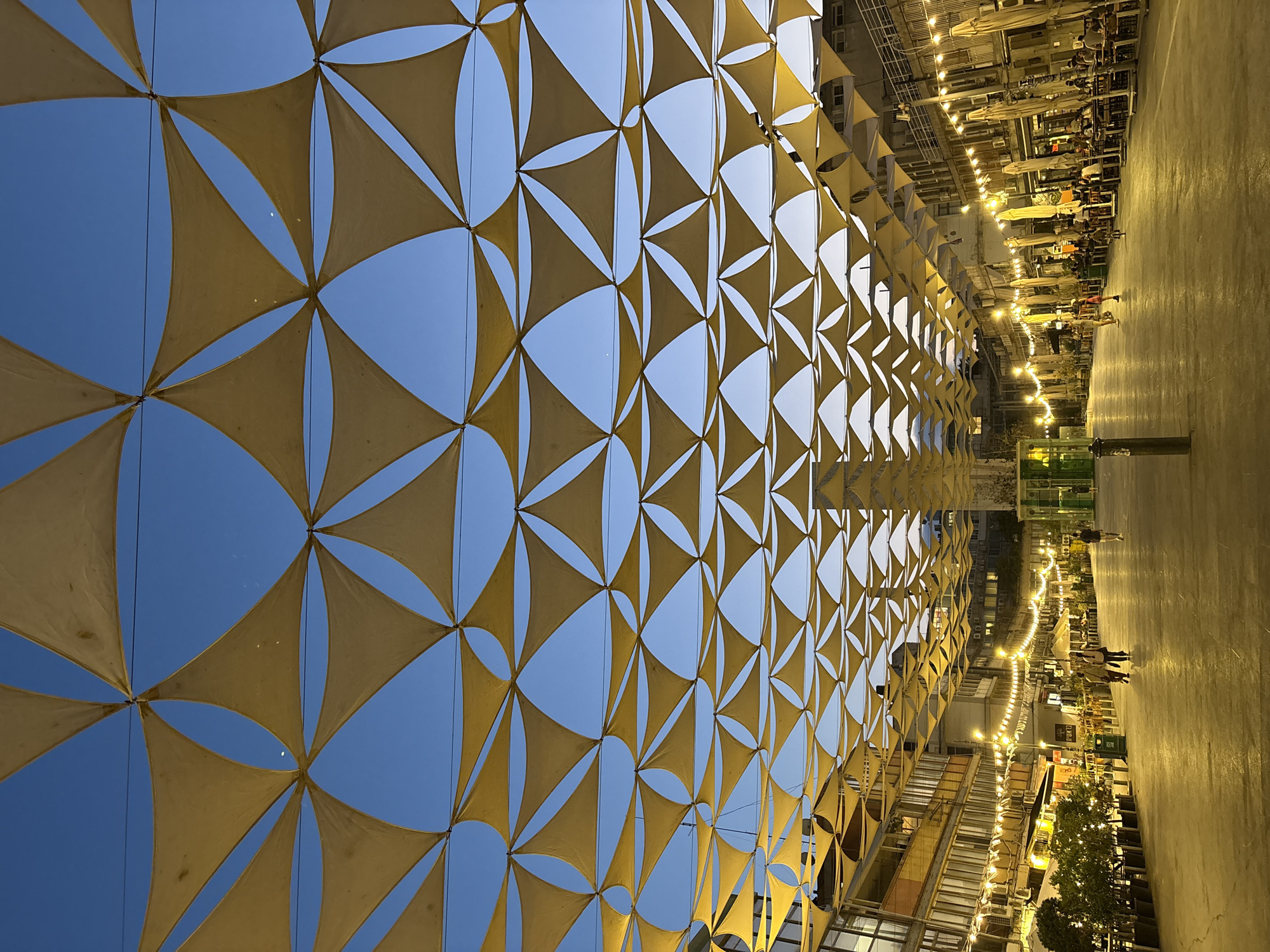 The image shows a street scene in Tel Aviv, Israel, at dusk. The street is lined with buildings on both sides, and a large canopy of yellow triangular fabric shades is stretched overhead, creating a geometric pattern against the twilight sky. String lights are hung along the buildings, illuminating the pedestrian walkway and outdoor seating areas of restaurants. A few people are visible walking and sitting at tables. The overall impression is one of a lively, modern urban environment.