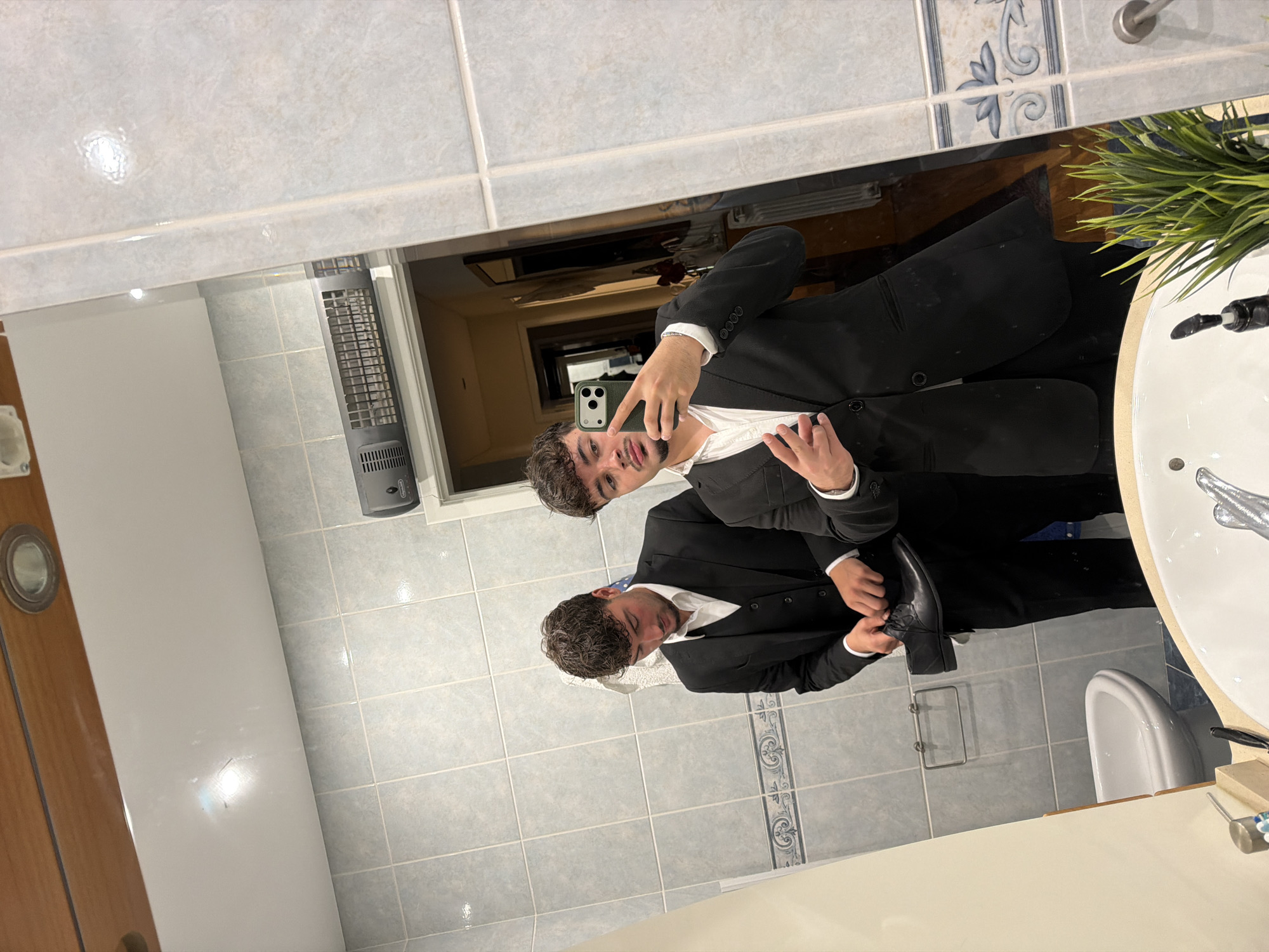 A reflection in a bathroom mirror shows two men in black suits, one holding a phone, possibly taking a selfie. The bathroom features light blue tiled walls, a white sink, and a toilet. A plant sits on the sink countertop. The interior of another room is visible behind the men in the reflection, suggesting a hallway or doorway. The specific location in Vila Real, Portugal cannot be determined from the image.