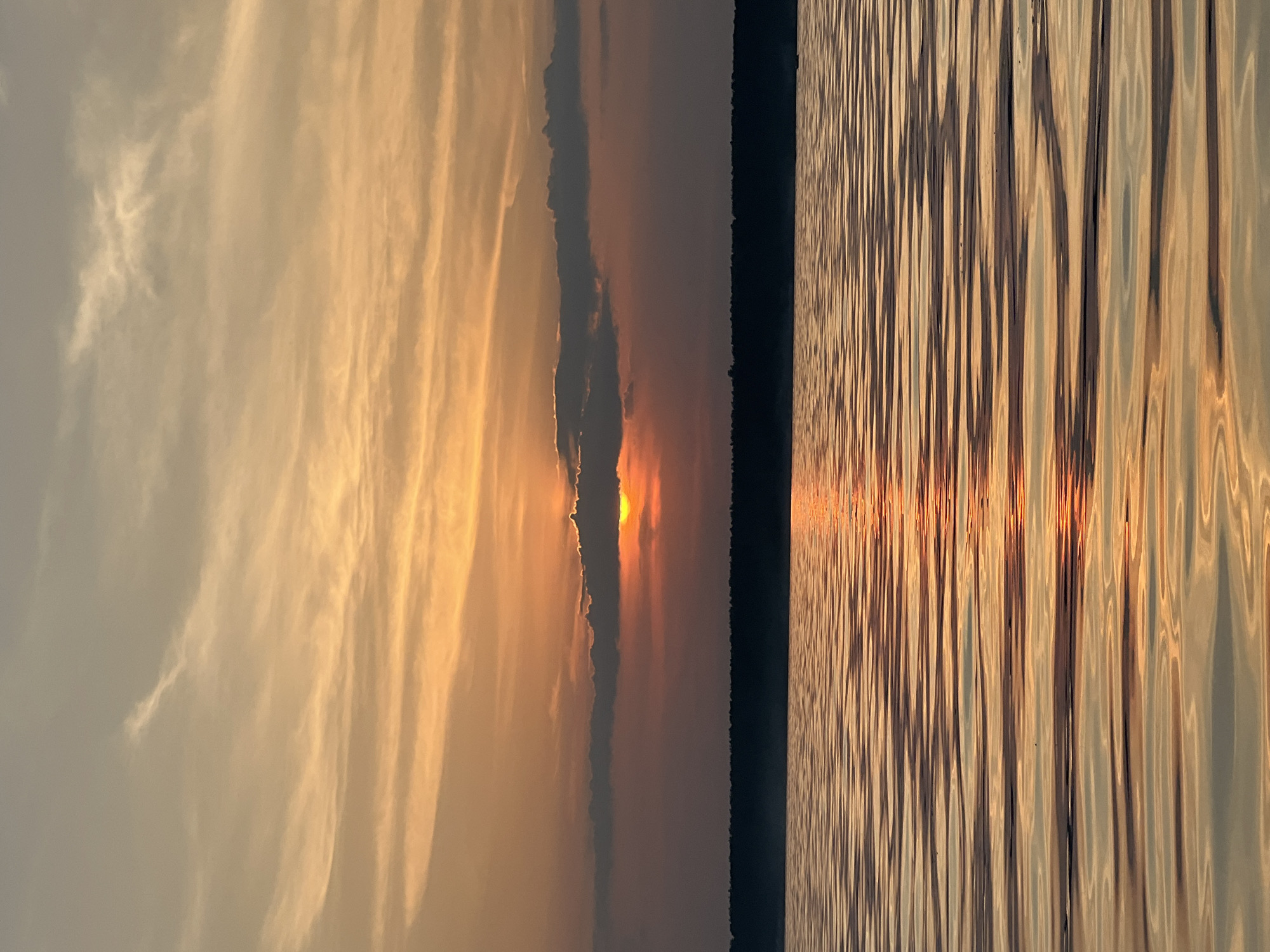The image depicts a serene sunset over a body of water, likely a lake, in Borovoye, Russia. The sky is a canvas of warm hues, transitioning from soft oranges and yellows near the horizon to a lighter beige at the top, with wispy clouds streaked across. A sliver of the sun is visible peeking through a horizontal band of darker clouds just above the tree line. The tree line itself appears as a dark silhouette, indicating a dense forest bordering the water. The water surface is calm, reflecting the colors of the sky and the sun, creating shimmering, rippling patterns of light and shadow. The overall scene evokes a sense of tranquility and natural beauty.