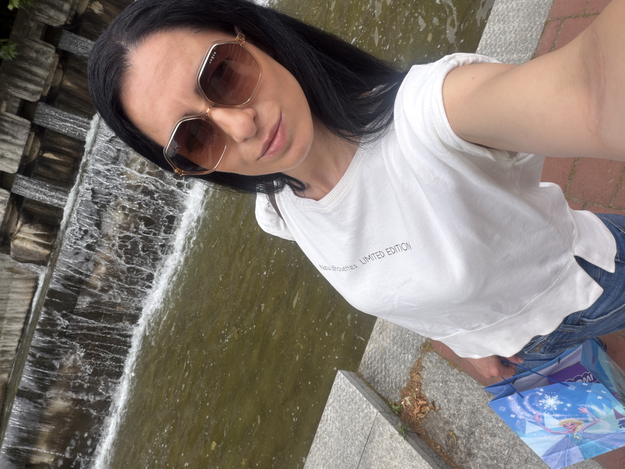 The image is a selfie of a woman with dark hair wearing large brown sunglasses and a white t-shirt with the words "New silhouettes LIMITED EDITION" printed on it. She is standing near a stone ledge with a waterfall in the background. The water appears to be slightly murky. To her right and below her are blue shopping bags featuring Disney's "Frozen" characters. The ledge is made of light gray stone with some debris and sparse plant growth visible. The woman is looking directly at the camera. The location is the artificial waterfall and water feature in Kaylaka Park, Pleven, Bulgaria.