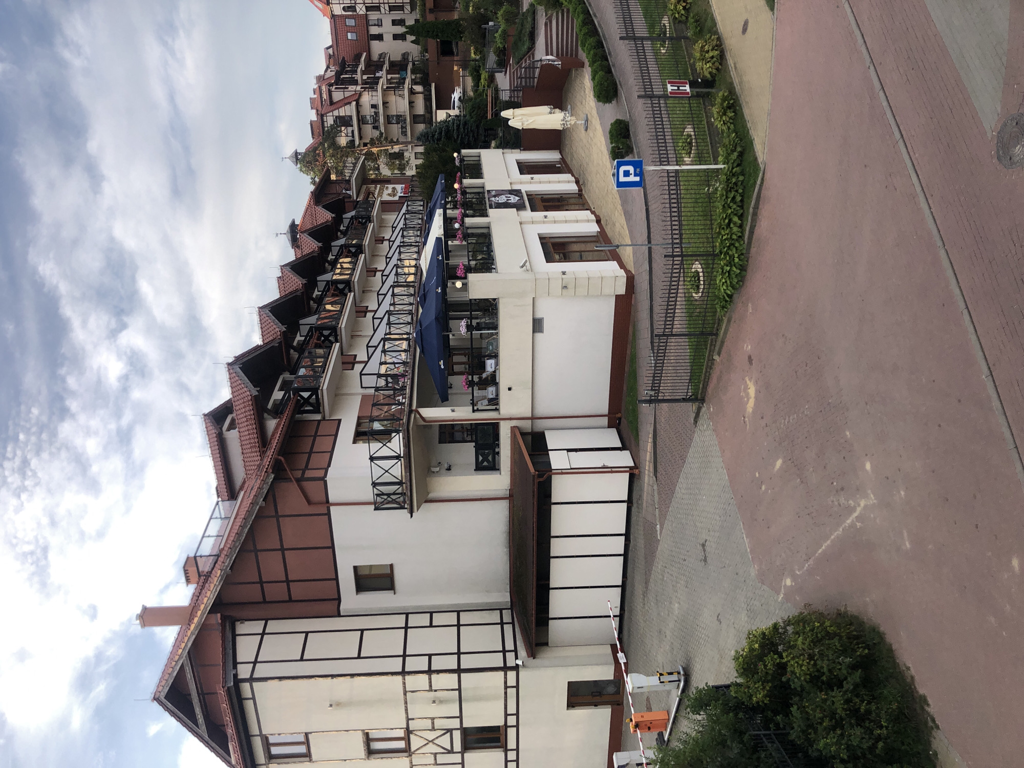 The image shows a multi-story building with a red-tiled, gabled roof. The building's facade is white with dark brown timber framing, giving it a traditional, almost Bavarian architectural style. It has multiple balconies with dark railings, and several windows of various sizes. In front of the building, there is an outdoor dining area with tables, chairs, and large blue umbrellas. A parking barrier is visible on the left side of the image, with a paved driveway leading up to it. A blue parking sign with a white "P" is posted near the building. The environment appears to be a resort or hotel complex, with well-maintained greenery and walkways. The sky is overcast with visible clouds.