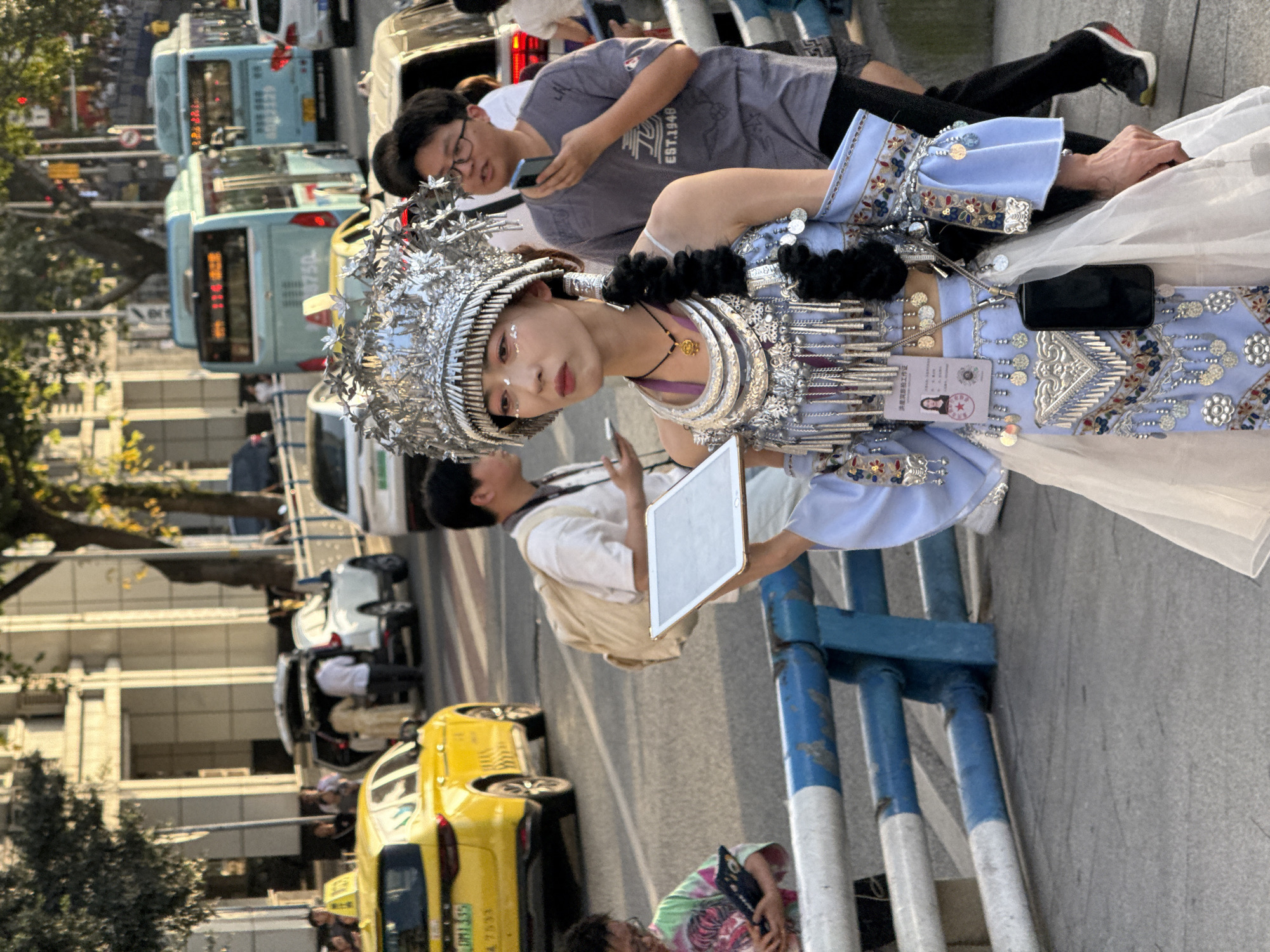 A medium shot depicts a person adorned in a silver headdress and traditional light blue and silver dress, standing on a city street. The person is looking directly at the camera, holding a white tablet. Behind the person, a crowd of people can be seen, some holding mobile phones, suggesting they may be taking photos. In the background, there is a busy street with several cars and buses. The scene is in Yuzhong District, China.