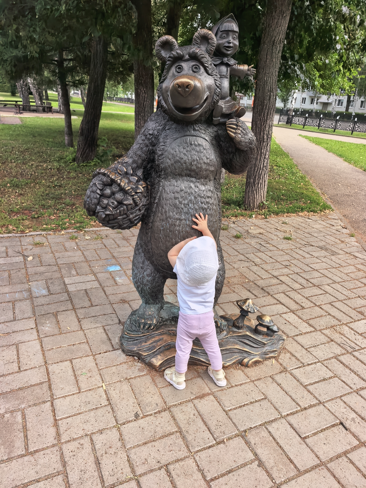 A bronze statue of a bear carrying a child on its shoulders stands in a park in Sterlitamak, Russia. The bear is depicted holding a basket of berries in its left paw. A young child, wearing a white hat, t-shirt, and pink leggings, stands facing the statue with their right hand reaching out towards the bear. The statue is situated on a paved walkway, surrounded by trees and a grassy area. In the background, there is a fence, buildings, and more trees. The lighting suggests it is daytime.
