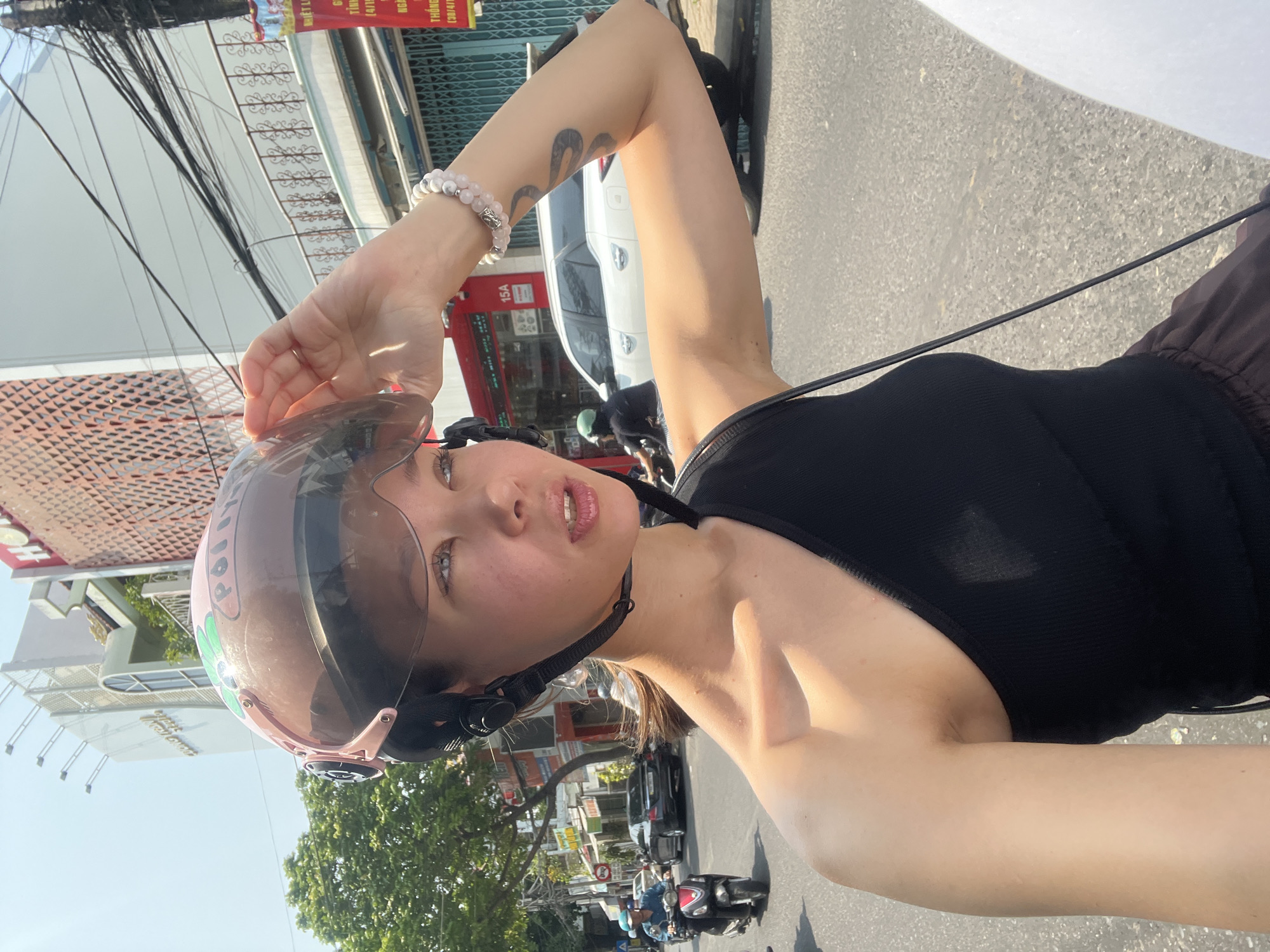 A woman is taking a selfie while riding a motorcycle on a street in Nha Trang, Vietnam. She is wearing a black top and a pink helmet with the word "POLICE" printed on it. She has a tattoo on her right arm and a bracelet on her left wrist. In the background, there is a street with cars, motorcycles, buildings, and overhead power lines. One building, marked 15A, displays signage with Vietnamese text.