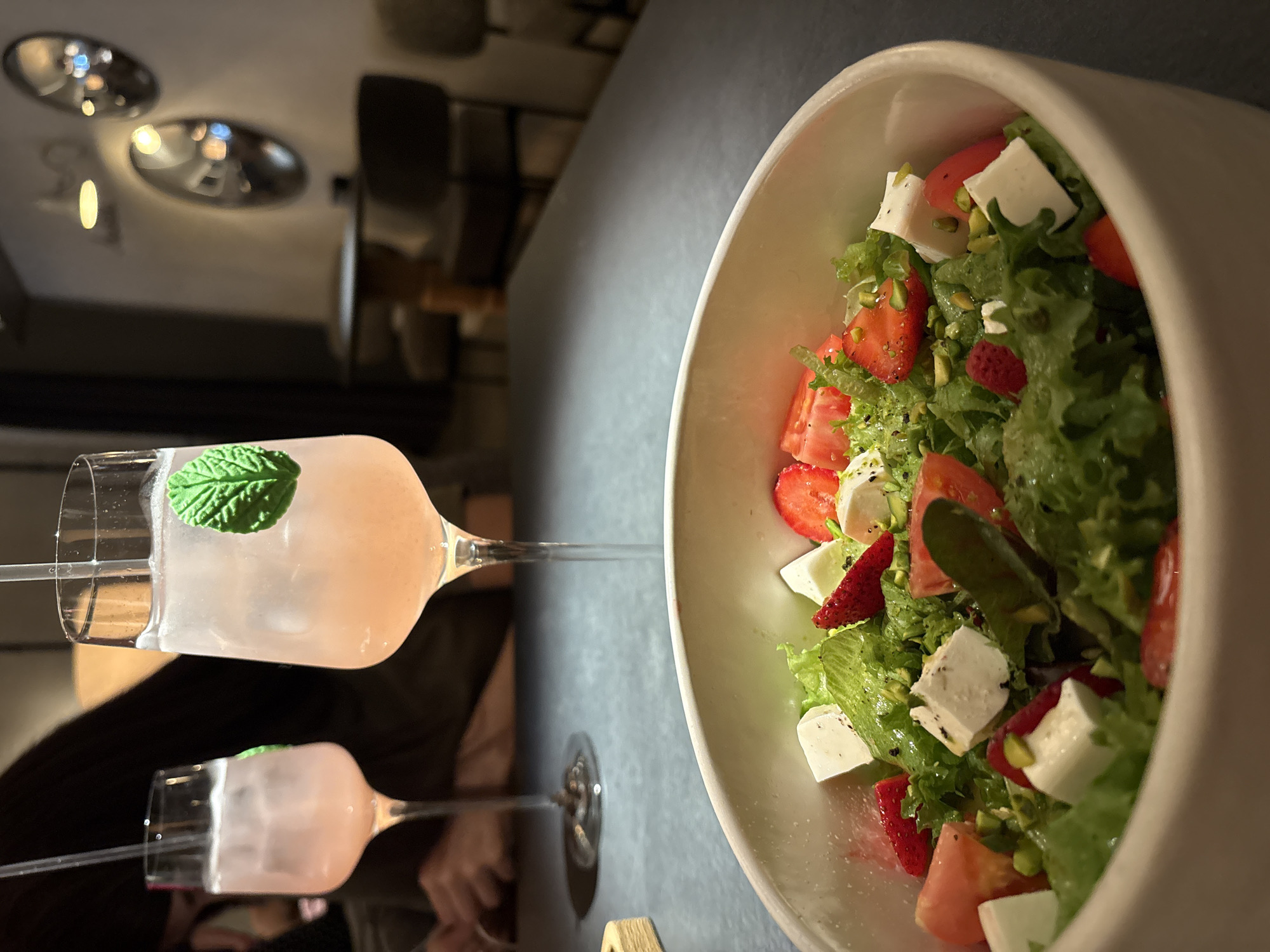 The image captures a meal setting, with a glass of pink lemonade garnished with a mint leaf and a large bowl of salad as the main focus. The salad appears to be composed of lettuce, sliced strawberries, diced tomatoes, and feta cheese, possibly with a sprinkle of nuts. Both items are resting on a dark grey tabletop, which shows a reflection of the lemonade glass. The background is softly blurred, hinting at an indoor dining environment, with hints of seating and muted lighting. The location is potentially a restaurant or cafe, in Krasnoyarsk, Russia.
