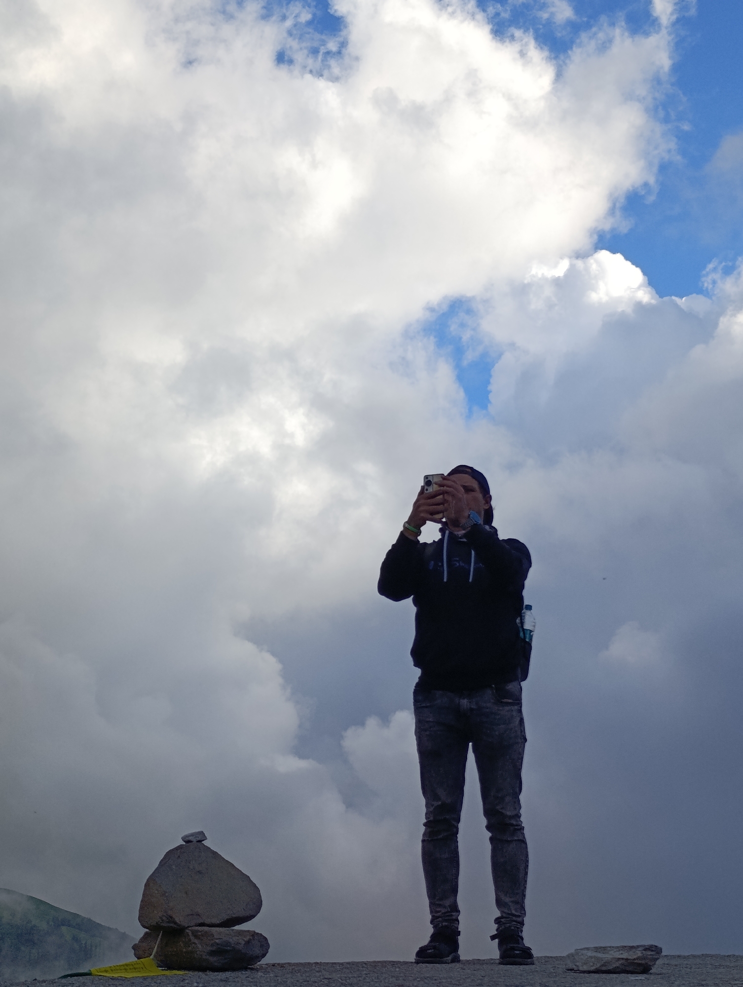 In Marhi, India, a person stands on a rocky ledge, silhouetted against a dramatic sky filled with white and grey clouds. They are wearing a dark hooded sweatshirt, dark jeans, and a baseball cap, and are holding a smartphone up to take a photograph. Their posture suggests they are focusing on capturing the expansive cloudscape above. In the foreground, a small cairn made of stacked stones stands to the left of the person. The ground appears to be gravelly or rocky. To the far left, a mountain slope is visible, partially obscured by mist. The overall impression is one of a person experiencing the grandeur of the natural environment in a remote, high-altitude location.