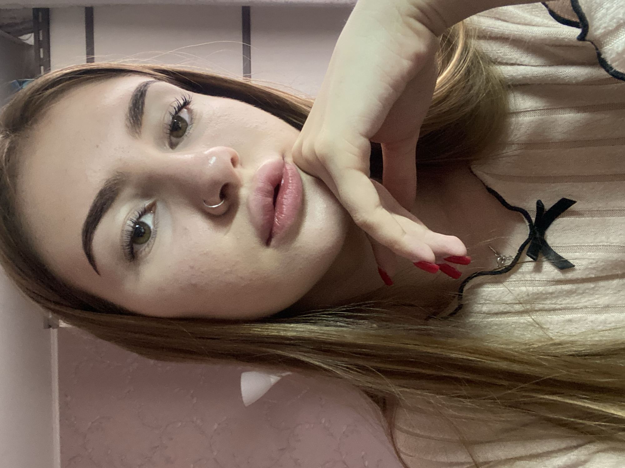 A young woman with long, straight blonde hair and a nose ring is pictured in a close-up shot. She is wearing a light-colored ribbed top with black trim and a black bow detail at the neckline. Her right hand is resting on her chin, with bright red painted fingernails. The background appears to be an indoor setting with a textured pink wall and some indistinct shelving or furniture. The image does not provide any information about the location being in Дьяково, Russia, or any other specific geographical context.