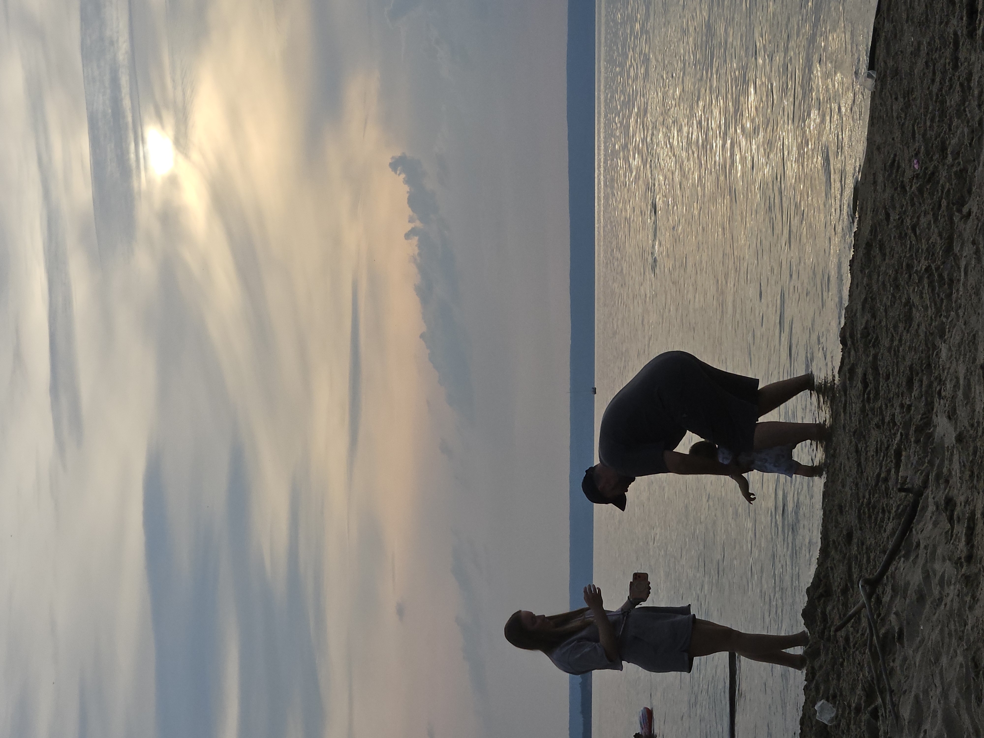 In the city of Leninsky, Russia, at sunset, a family is enjoying an evening by the water. A woman stands on the sandy shore, holding what appears to be a phone and a cup, facing a man who is partially in the water. The man is bent over, interacting with a young child who is also in the shallow water. The child is facing away from the camera, reaching out a hand. The sun is low in the sky, casting a warm glow and reflecting on the rippling surface of the water. The background shows a distant shoreline and a sky filled with wispy clouds.