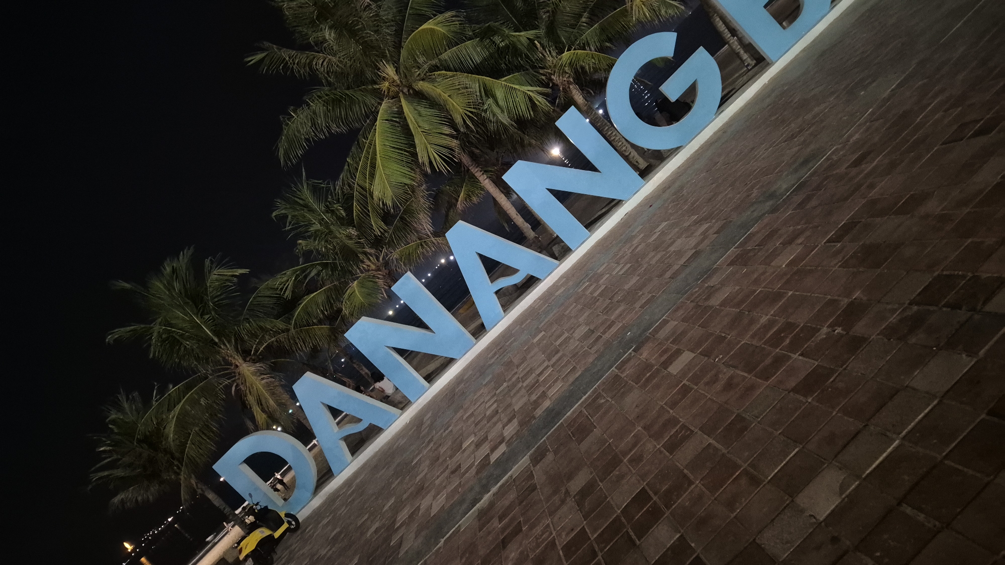 A large, light blue sign spelling out "DA NANG" is displayed diagonally across a brick-tiled walkway. The letters are illuminated, creating a soft glow against the dark night sky. Palm trees with green fronds frame the sign, their silhouettes visible against the night. In the background, faint lights suggest a city or waterfront area. To the left of the sign, a yellow electric scooter is parked. The scene appears to be a public area, possibly a promenade or park, in the city of Da Nang, Vietnam.