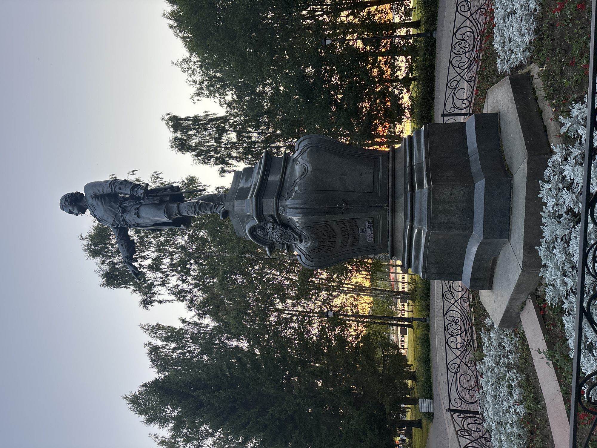 A dark bronze statue of Peter the Great stands in Petrozavodsk, Russia, against a pale, clear sky. The statue depicts Peter the Great in a military uniform, with his left arm extended, pointing forward, and his right hand on the hilt of his sword. He is standing on a tall, ornate pedestal, also made of dark bronze, which has inscriptions on its sides. The pedestal is surrounded by a low black metal fence with intricate scrollwork. In front of the fence, there are beds of low-lying plants with silvery-grey leaves and some red flowers. Behind the statue and fence, a line of tall, slender trees forms a dense backdrop. The trees are a mix of evergreens and deciduous trees, with some of the deciduous trees showing golden light filtering through their leaves, suggesting it is either sunrise or sunset. A lamppost is visible among the trees. In the background, beyond the trees, there are hints of buildings and possibly a body of water, with more trees and structures visible further in the distance. The ground in front of the trees appears to be a grassy area with pathways.