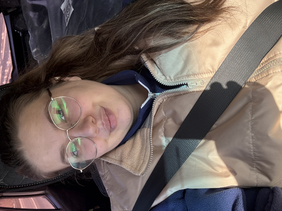 A young adult female is depicted seated in the passenger side of a vehicle in the city of Kukushtan, Russia. She has light skin, long brown hair, and is wearing round, metallic-rimmed glasses. Her facial expression shows a slight smile with pursed lips, and she looks directly towards the camera. She wears a light beige or tan puffer vest over a dark blue long-sleeved top with a light blue zipper. A dark grey seatbelt crosses her chest diagonally from her left shoulder to her right hip. The vehicle's interior is visible, including a dark headrest behind her and an out-of-focus window showing a blurred exterior scene to her left. Natural light illuminates her from the left side, creating a shadow across her chest from the seatbelt.