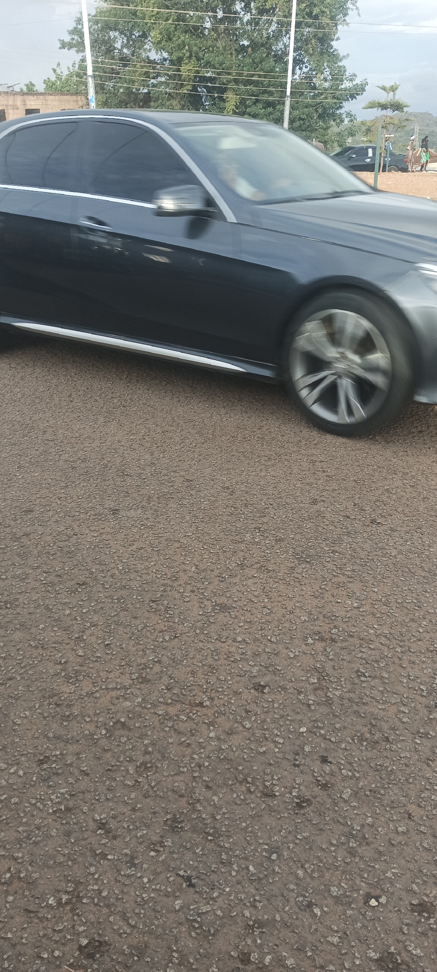 The image depicts a dark-colored sedan, likely a Mercedes-Benz, in motion from left to right across the frame. The vehicle features tinted windows and a silver trim strip along the lower side. Its front right wheel exhibits motion blur, indicating movement. The road surface in the foreground and beneath the car is composed of reddish-brown gravel or coarse asphalt with scattered small stones.
In the midground and background, various elements are visible. To the left, there are dense green trees and a section of a light-colored building with a flat roof. Utility poles and overhead lines are present across the upper mid-section. Towards the right background, a dark-colored pickup truck is partially visible, appearing to be parked or stationary. Several individuals are standing or walking in the far right background, near more trees and what appears to be a distant rocky hill or mountainous terrain. The scene is illuminated by daylight, suggesting an outdoor setting in the city of Bukuru, Nigeria.