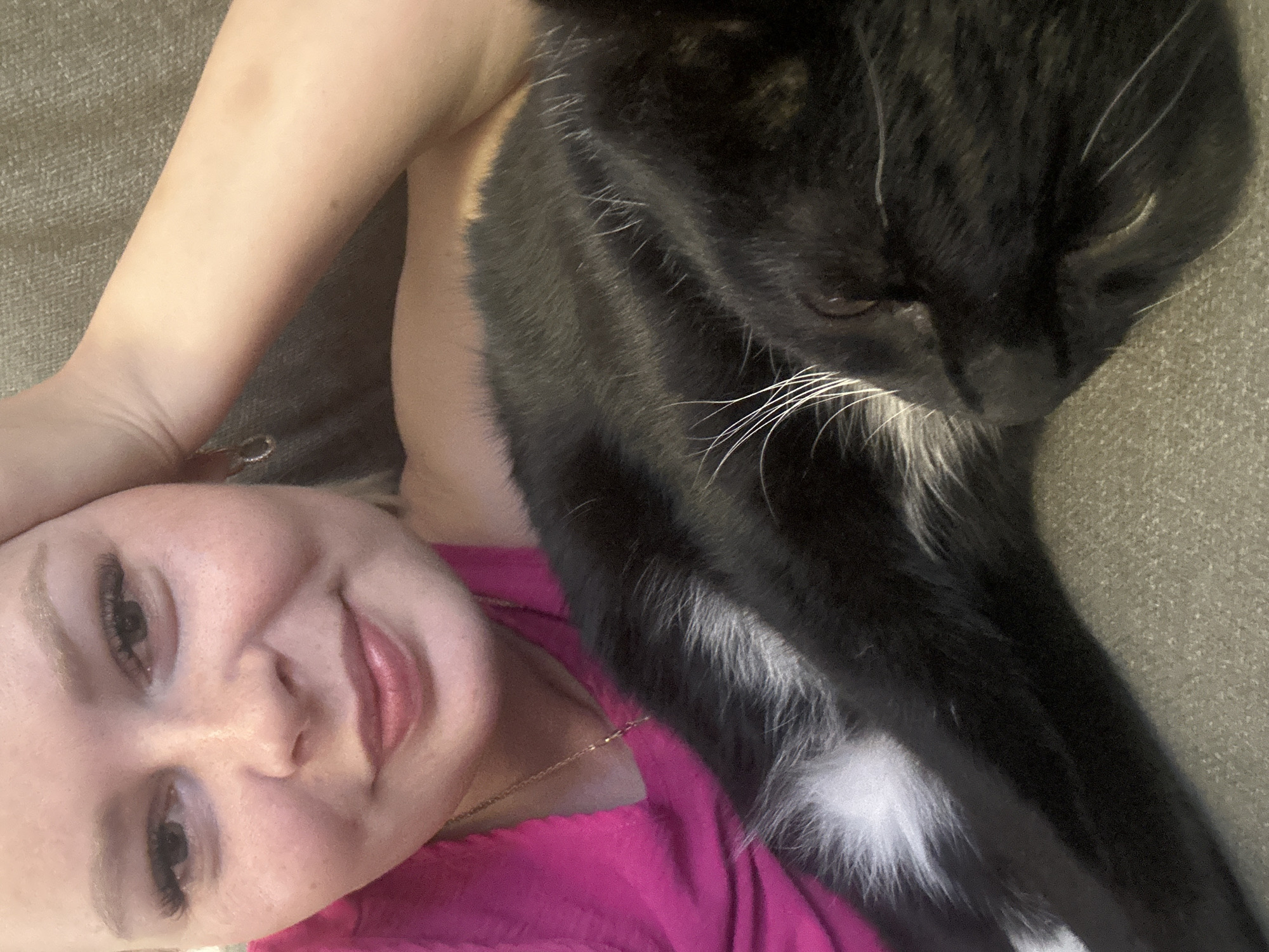 A close-up selfie shows a light-skinned person with blonde hair and heavy eyeliner resting their head on their arm, with a black cat snuggled next to them. The person is wearing a bright pink top and a delicate necklace. The cat is mostly black with some white markings on its chest. They are both lying on a textured, neutral-colored surface, possibly a sofa. The background is slightly out of focus, but appears to be an indoor setting. There are no discernible elements in the image that can place the location in Abakan, Russia.