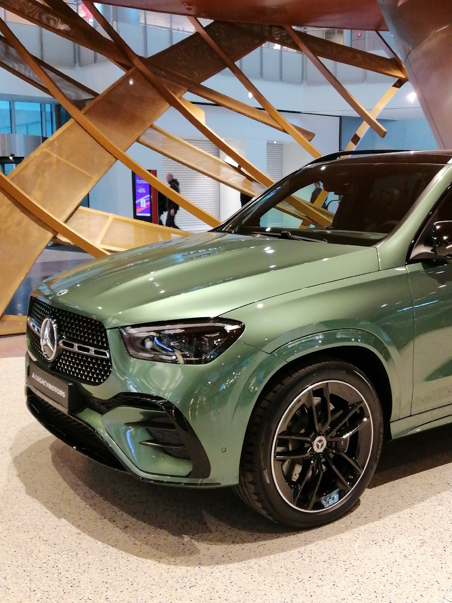The image shows a green Mercedes-Benz GLE parked indoors, likely in a showroom or exhibition space. The car is the primary subject, with a focus on its front and driver's side. The background features a modern, artistic wooden structure with intersecting beams. In the background, there are faint indications of people, one of which is by a monitor displaying a "Family" and "-85%" sign. The location is identified as Moscow, Russia. The lighting is bright and even, highlighting the car's features and color. The floor has a speckled, terrazzo-like appearance. A "A LEGACYMOTORS" license plate is visible.