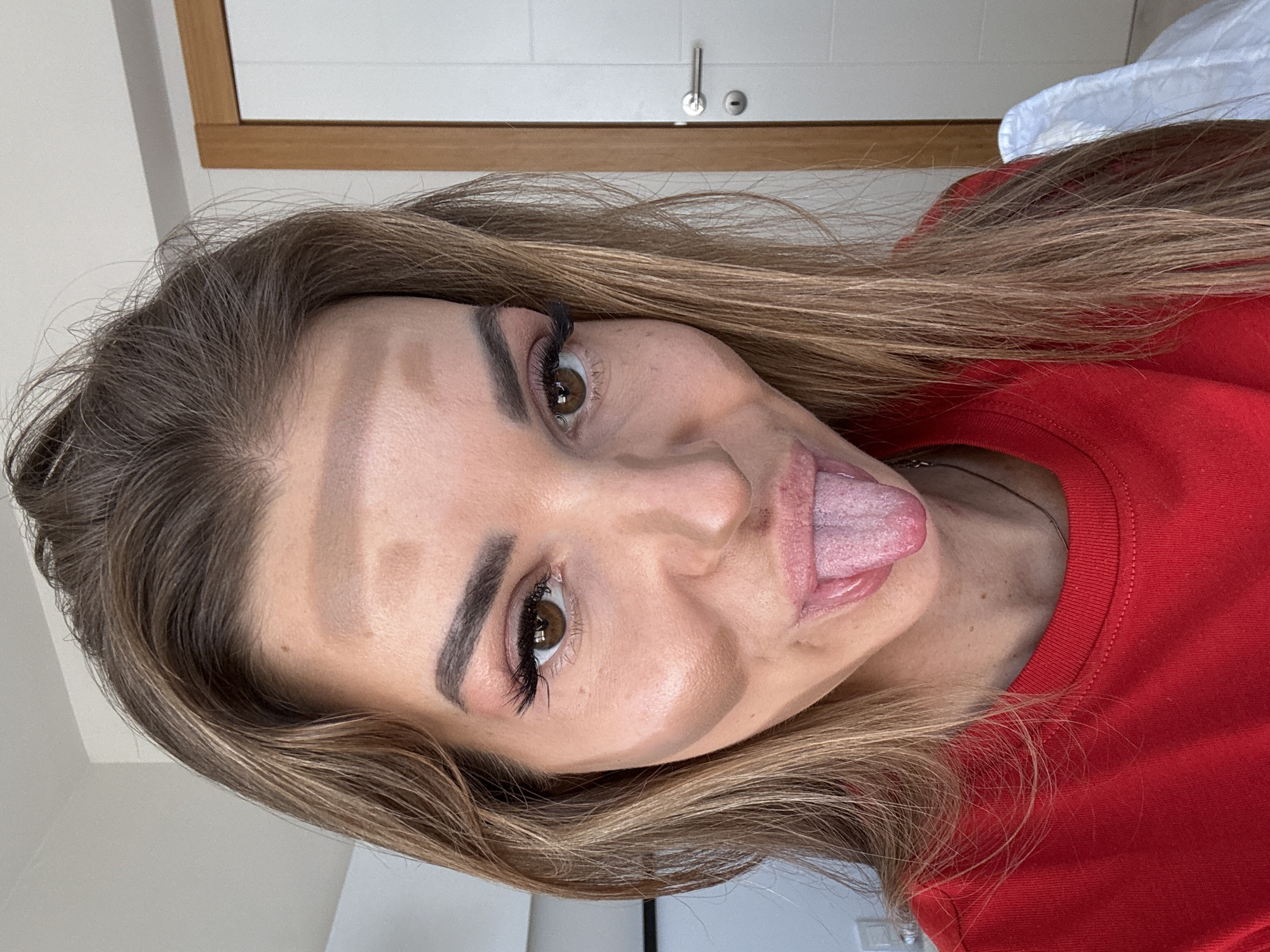 A woman with long brown hair and brown eyes is close-up, sticking out her tongue, wearing a red shirt. The background shows a white door with a silver handle, and a wooden frame around the door. The environment appears to be indoors. The location is not discernible from the image.