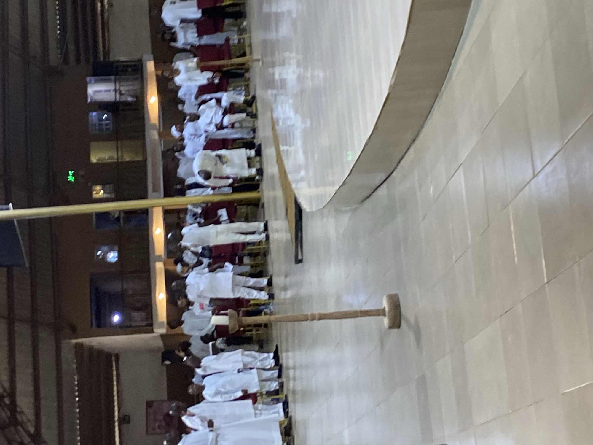 The image depicts a gathering of people in what appears to be a religious or ceremonial setting, possibly in Rukuba Road, Nigeria. The individuals are dressed in predominantly white garments, with some wearing maroon or red accents. They are arranged in rows, suggesting an organized assembly. A large wooden mallet-like object is visible on the floor, oriented lengthwise toward the group. To the side, a curved structure is present, possibly a stage or architectural element. In the background, a building can be seen with windows and what appears to be an electronic display. The flooring is tiled and reflective, indicating a polished surface. The scene is captured under artificial lighting, casting shadows across the room.