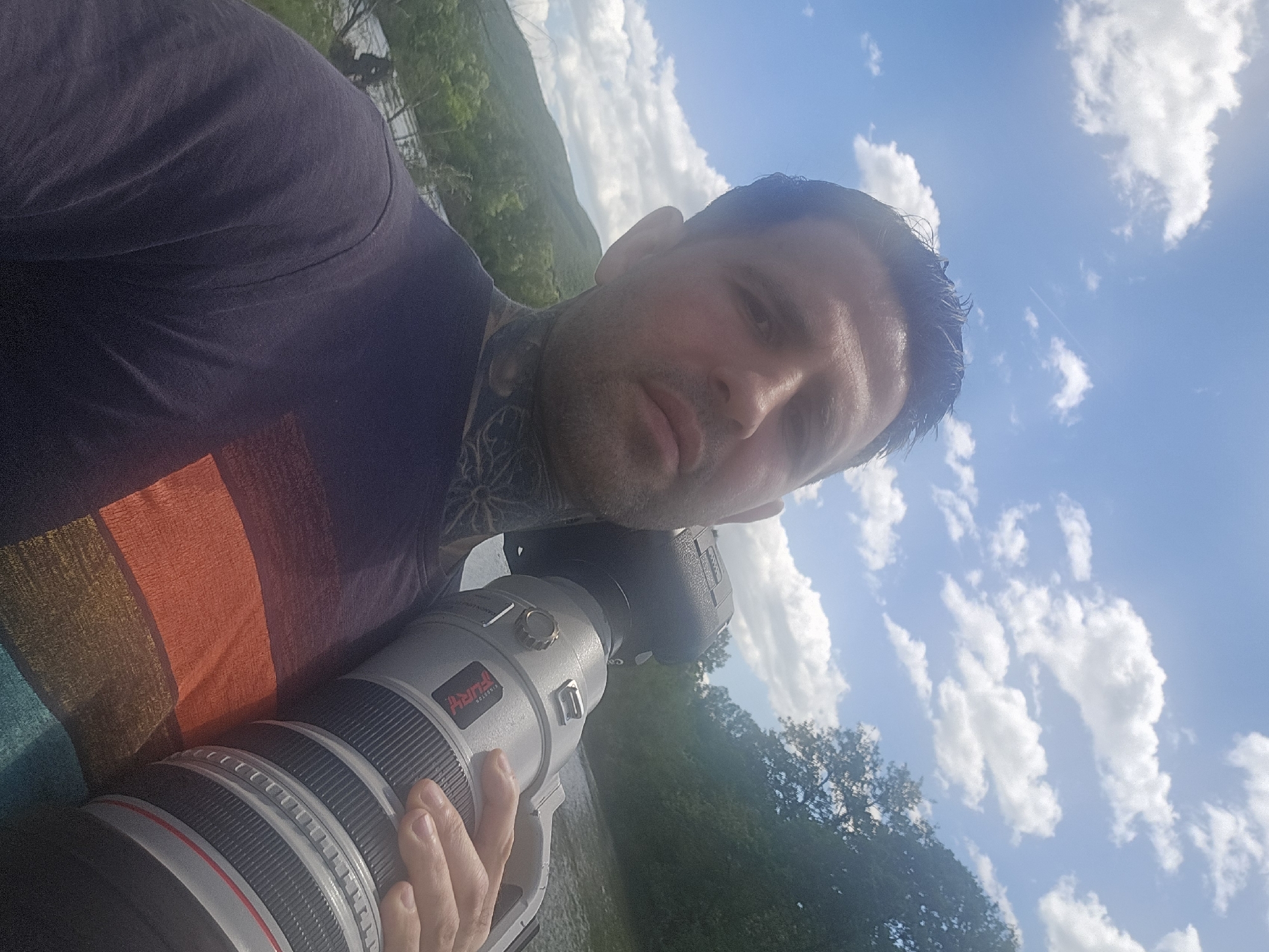 A selfie taken outdoors in Zimnitsa, Bulgaria, shows a man holding a professional camera with a large telephoto lens. The man has short dark hair and tattoos on his neck. He is wearing a t-shirt with orange, brown, and blue stripes. He is positioned with a view of green hills and trees in the background. The sky is visible, with scattered white clouds. The photo angle is tilted.