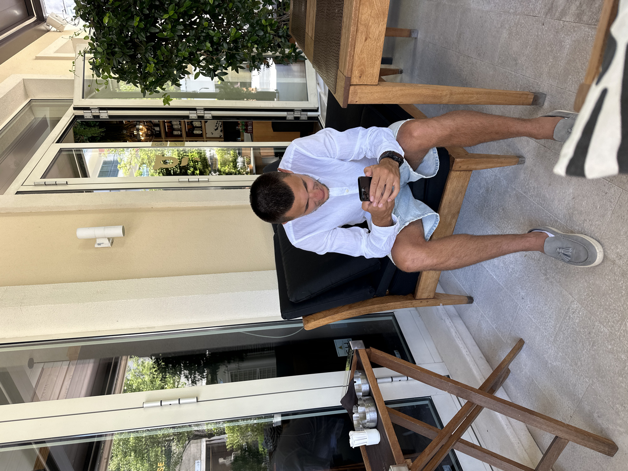 A young man with short dark hair is sitting on a black chair on an outdoor patio, looking at his smartphone. He is wearing a white button-down shirt and light blue denim shorts. He has on grey tassel loafers and white socks. To his left, on a wooden folding table, are three ashtrays and a small white cup containing toothpicks. To his right is a wooden table with a dark textured placemat. In the background, there are large glass doors leading into a building, revealing shelves stocked with bottles. Green foliage is visible both inside the building and as a large bush to the right of the man. The patio floor is tiled with light-colored stone. The scene appears to be located at a restaurant or cafe in the city of Tivat, Montenegro, based on a partially visible sign on the glass doors that reads "STEAKHO".