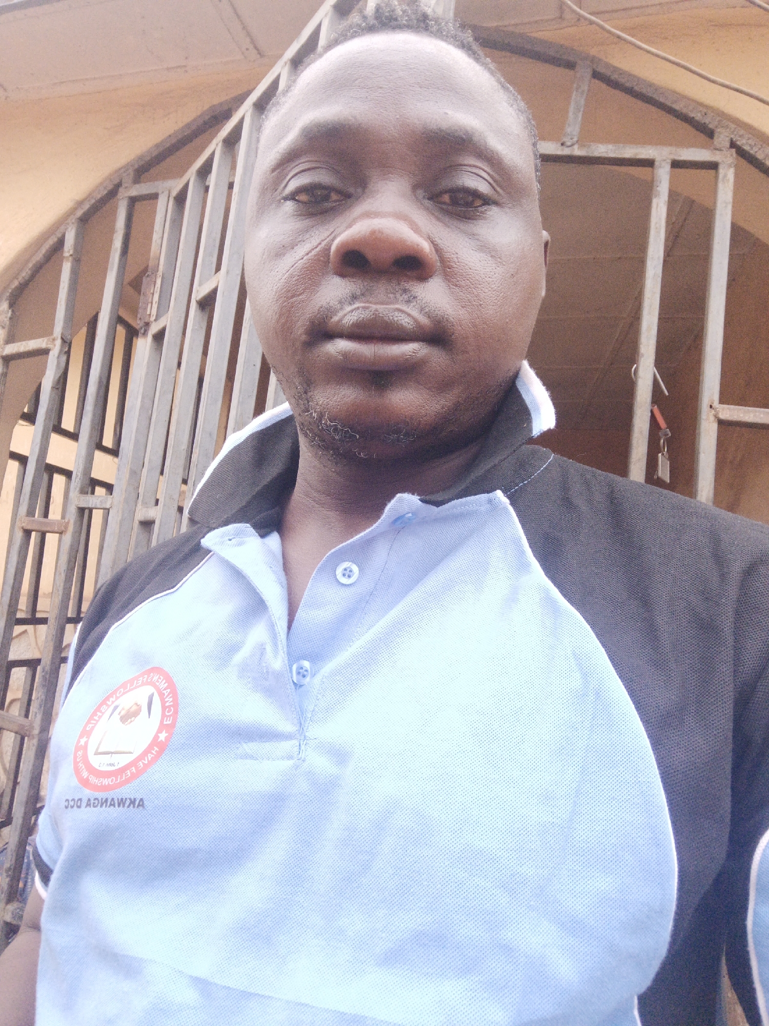 A medium close-up shot of a man facing the camera, taken during daylight hours in Akwanga, Nigeria. The man is wearing a two-tone polo shirt, with a light blue front and black sleeves. The shirt has a logo on the left side of his chest. The background consists of a metal gate and a building structure. A set of keys is hanging in the upper right background.