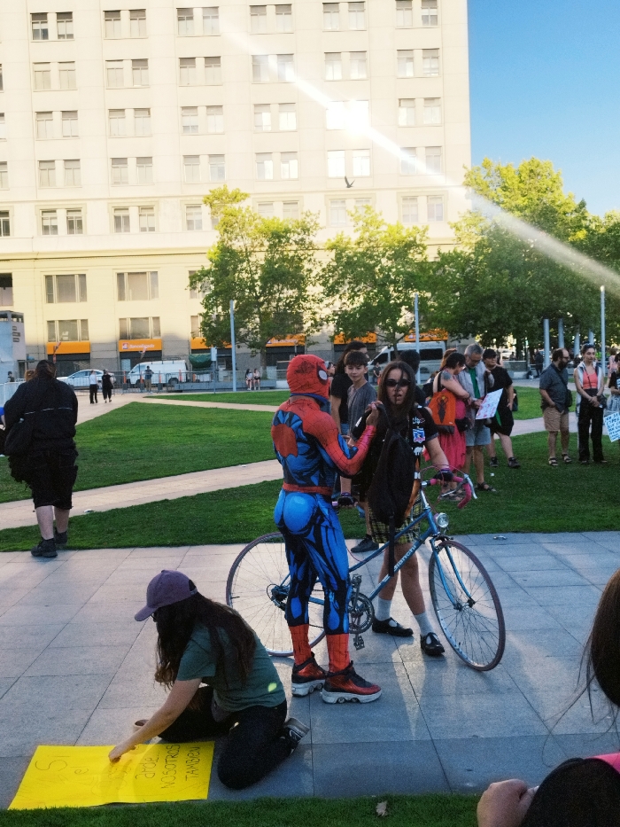 A person dressed as Spider-Man stands with a blue Bianchi bicycle. A girl wearing a plaid skirt stands with the bike as well. A building is in the background, and there are trees. A woman kneels on the ground drawing on a yellow poster. Several people stand in the background near the trees.
