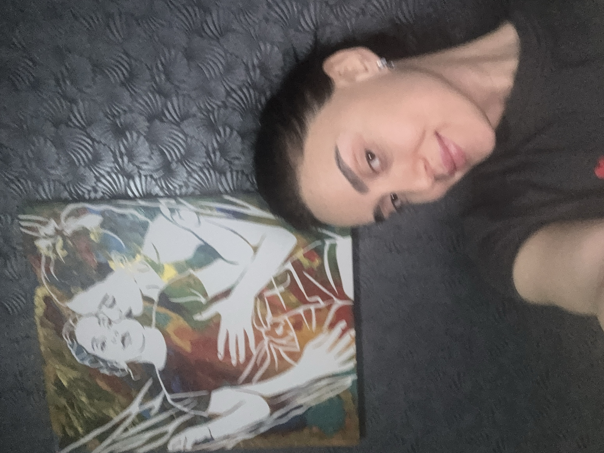 A woman is lying on a dark gray textured surface next to a square painting. The painting depicts two people in a figurative style with white figures on a background of mixed colors. The woman in the photo has dark hair and is wearing a dark gray shirt. The location appears to be indoors. There is no indication of the city of Tbilisi, Georgia.