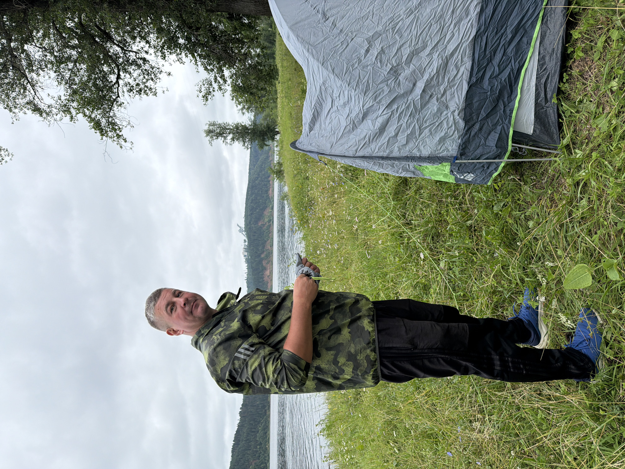 In Noviy, Russia, a man stands on a grassy riverbank next to a gray tent. He is wearing a camouflage jacket and black pants with blue sneakers. The man is holding a small bag in his hand. A wide river is behind him, with a forested hill on the opposite bank. The sky is overcast.