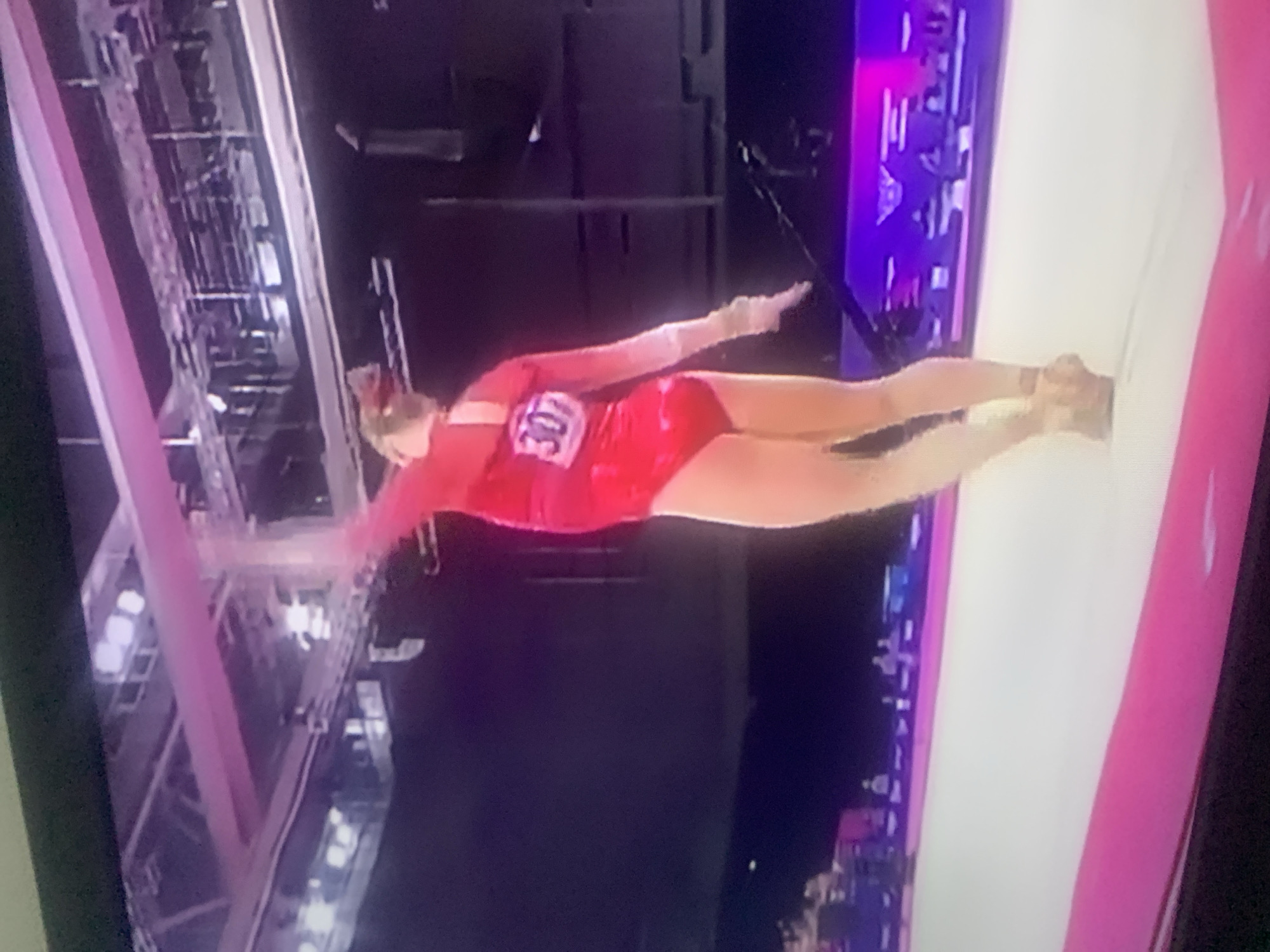 The image shows a gymnast in a red leotard with the number 307 on her back, performing on a white mat in an indoor arena. She is in mid-pose, with one arm extended upwards and the other outstretched to her side. The background is dark, with lights and what appear to be spectators in the distance. There is no indication of the city of Jos, Nigeria, or any specific location within it.