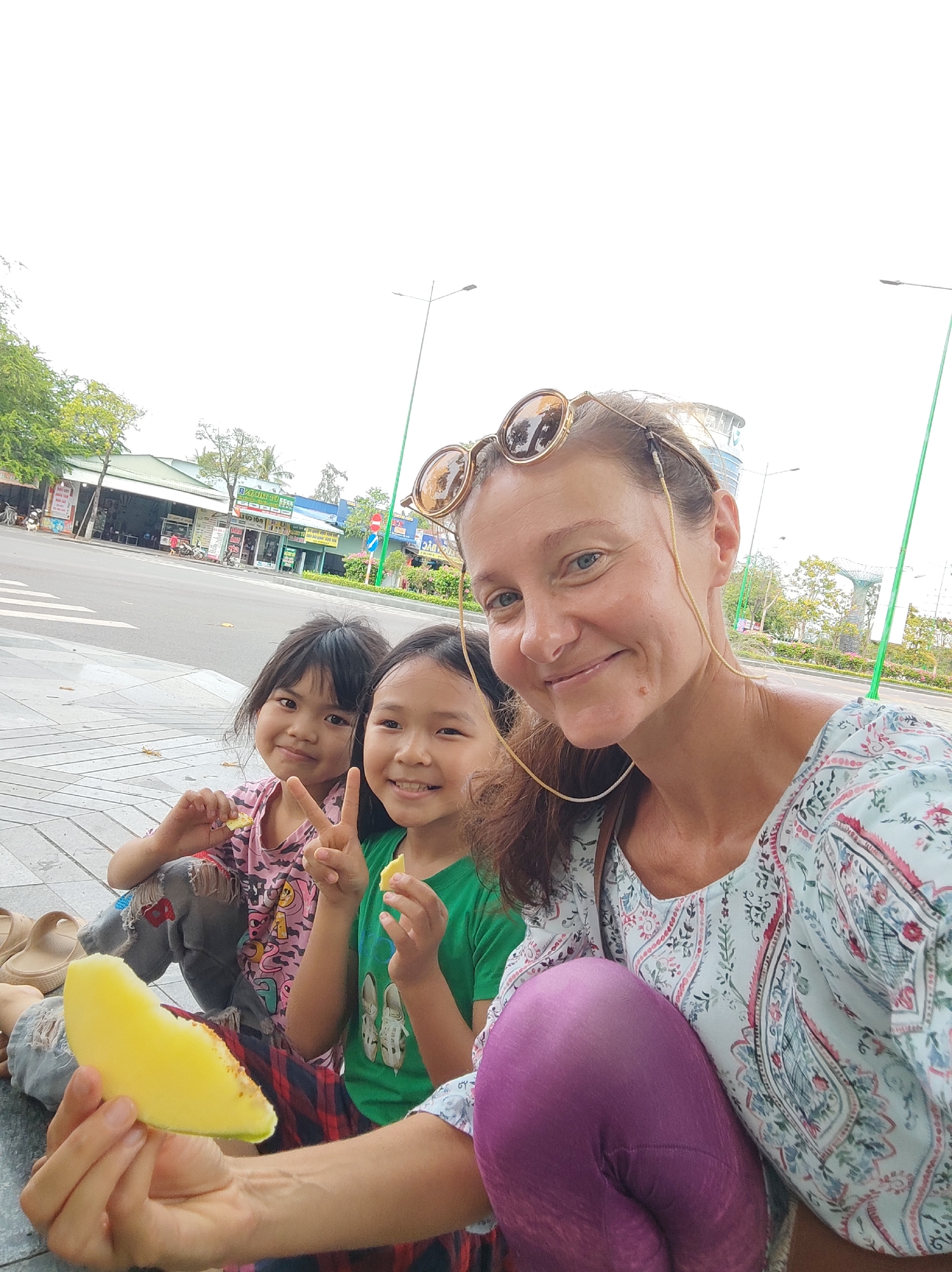 A woman with sunglasses perched on her head is taking a selfie with two young Asian girls on a city street. The woman is wearing a light-colored paisley blouse and purple leggings, and is holding a large piece of yellow fruit, possibly mango or melon. The girl on the left wears a pink and black animal-print shirt with distressed gray jeans, and holds a small piece of fruit. The girl on the right wears a green shirt with a white graphic and is giving a peace sign. They are sitting on a paved area adjacent to a street. In the background, there's a multi-lane road, buildings, trees, and streetlights.