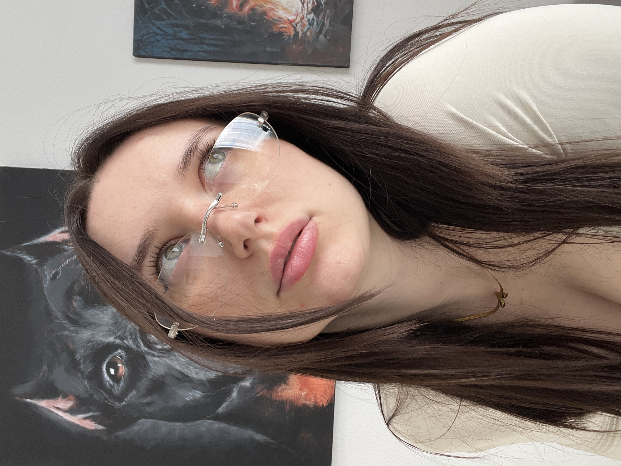 A close-up, rotated image shows a woman with long, dark brown hair, wearing frameless glasses, and a light-colored shirt. Her skin is fair, and she has light-colored eyes. Her lips are pink and appear to have lipstick on. She is wearing a gold necklace. To her left are two paintings. The painting closest to her face features a dark-colored dog. The scene appears to be indoors, and the background is blurred. There is no information in the image to indicate that the location is in the city of Tyumen, Russia.