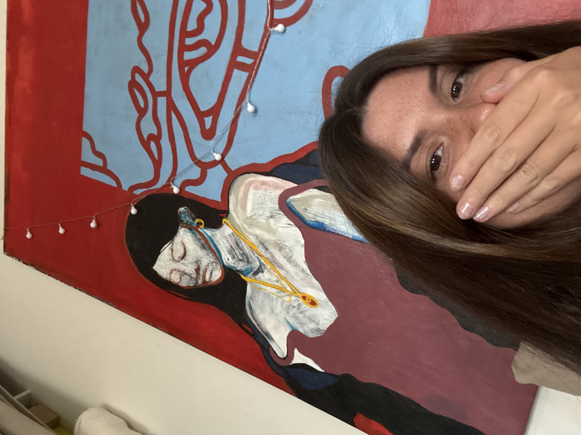 A close-up selfie captures a woman partially covering her mouth with her hand, looking towards the camera with her dark eyes. Her long, dark brown hair frames her face, revealing freckles on her nose and cheeks. Her fingernails are painted a light, natural shade.
The woman is positioned in an indoor setting, likely a room in Krasnoyarsk, Russia, in front of a vibrant painting that fills the background. The painting depicts a stylized figure with light skin and dark hair, appearing to be lying down, adorned with a distinctive gold-colored necklace or chain featuring an oval, red-accented pendant. The painting's background is characterized by a bold red section and a light blue section patterned with abstract, flowing red lines.
A string of small, white, spherical fairy lights is draped across the painting, adding a soft glow and decorative touch. The woman's gesture of covering her mouth, combined with her visible eye, suggests an emotion that could be shyness, surprise, or playfulness. The time of day is not discernible, as the scene is illuminated by indoor lighting. No text is visible in the media file.
