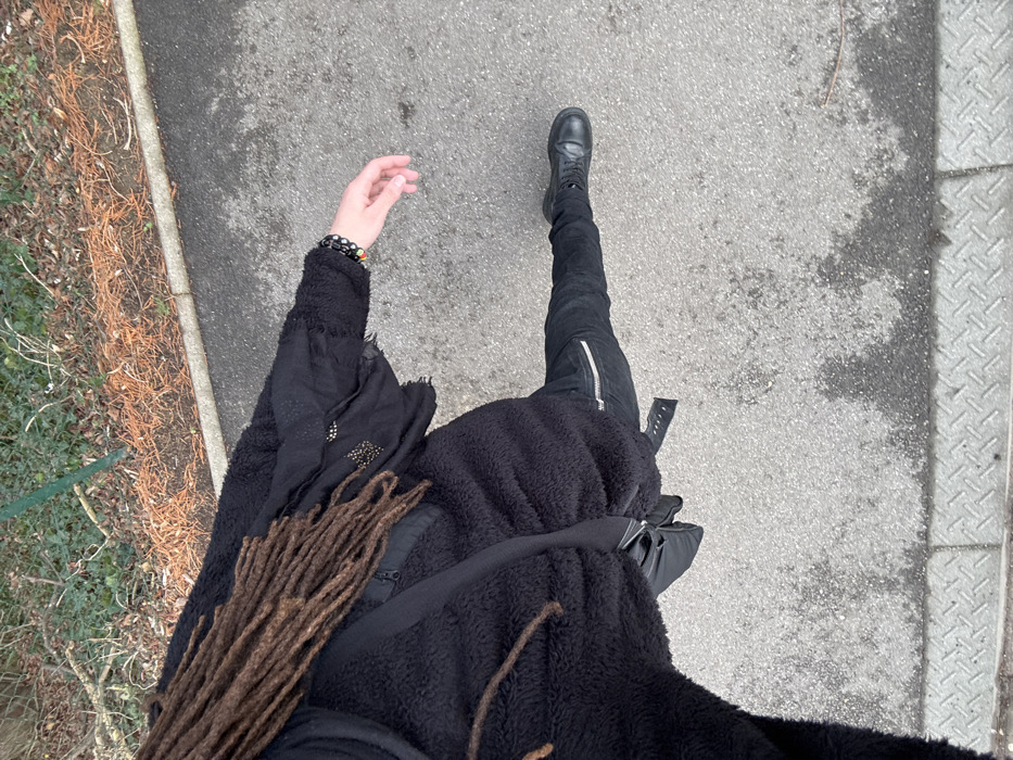 A subject is depicted from an overhead perspective, walking on a paved surface in Orsay, France. The individual wears a black, textured, long-sleeved jacket over a dark garment with scattered golden embellishments, black fitted trousers featuring a silver zipper pocket on the right thigh, and black lace-up boots. Brown dreadlocks are visible across the left shoulder and chest. The right arm is extended, with the hand slightly open and palm down, revealing a collection of dark and multicolored beaded bracelets on the wrist. A black fanny pack is secured around the subject's midsection. The ground is an asphalt pathway, characterized by mottled grey and darker, moisture-affected areas. A textured metal grate and a concrete curb define the edge of the pathway in the foreground. Beyond the path, a strip of sparse vegetation with brown leaf litter and green shrubbery is present, bisected by a thin green metal pole.
