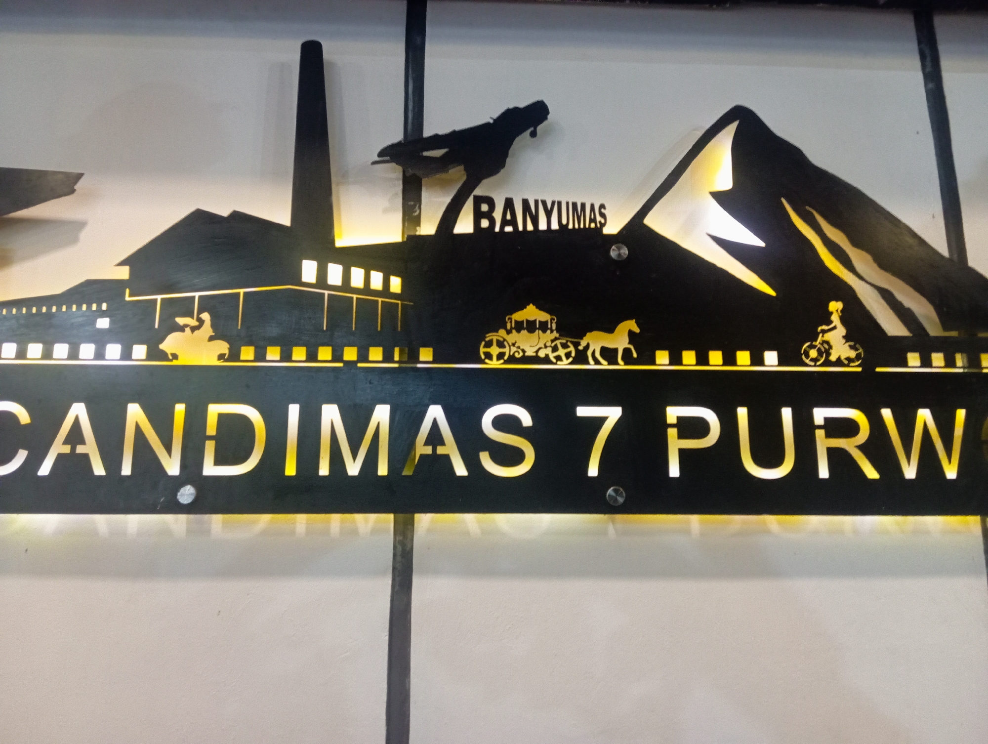 The image shows a sign with the text "CANDIMAS 7 PURW" illuminated by yellow light. The sign is a silhouette of a landscape with mountains, a factory chimney, a horse-drawn carriage, and a person on a bicycle. Above the landscape, the word "BANYUMAS" is visible. The sign appears to be mounted on a white wall. The overall scene is likely a decorative sign for a business or establishment in Purwanegara, Indonesia.